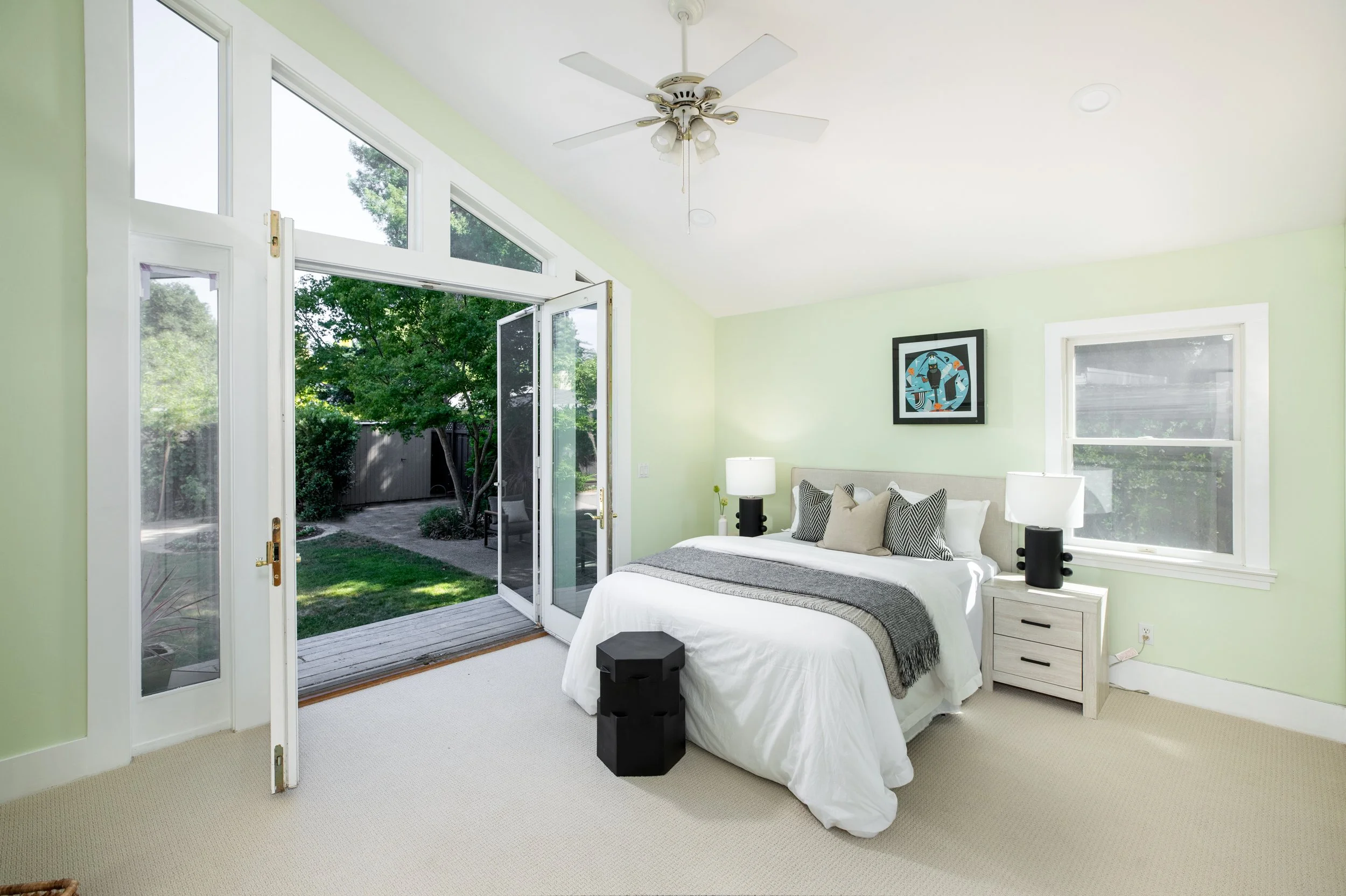 A bedroom with light green walls and a large window overlooking a backyard with trees. The bed has white bedding and striped pillows, with two black nightstands on either side and a white nightstand with a lamp on the right. A framed art piece is han