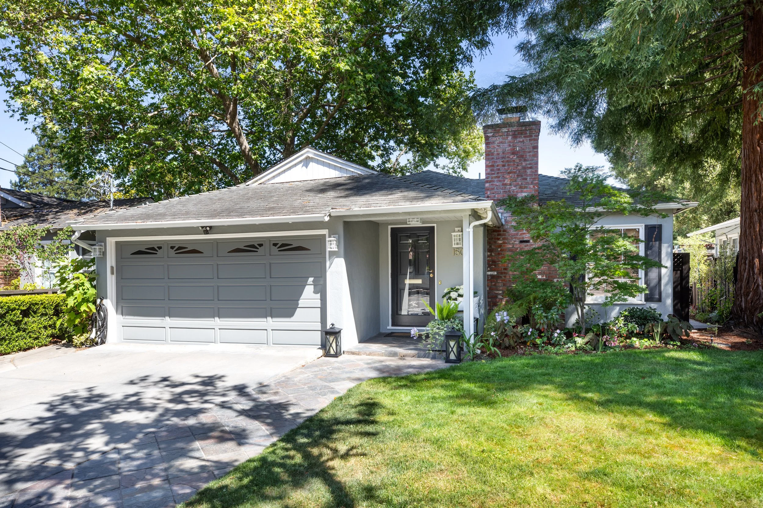 Front view of a house with a gray garage door, a black front door, and a chimney, surrounded by green trees and a well-maintained lawn with some potted plants and a stone pathway.
