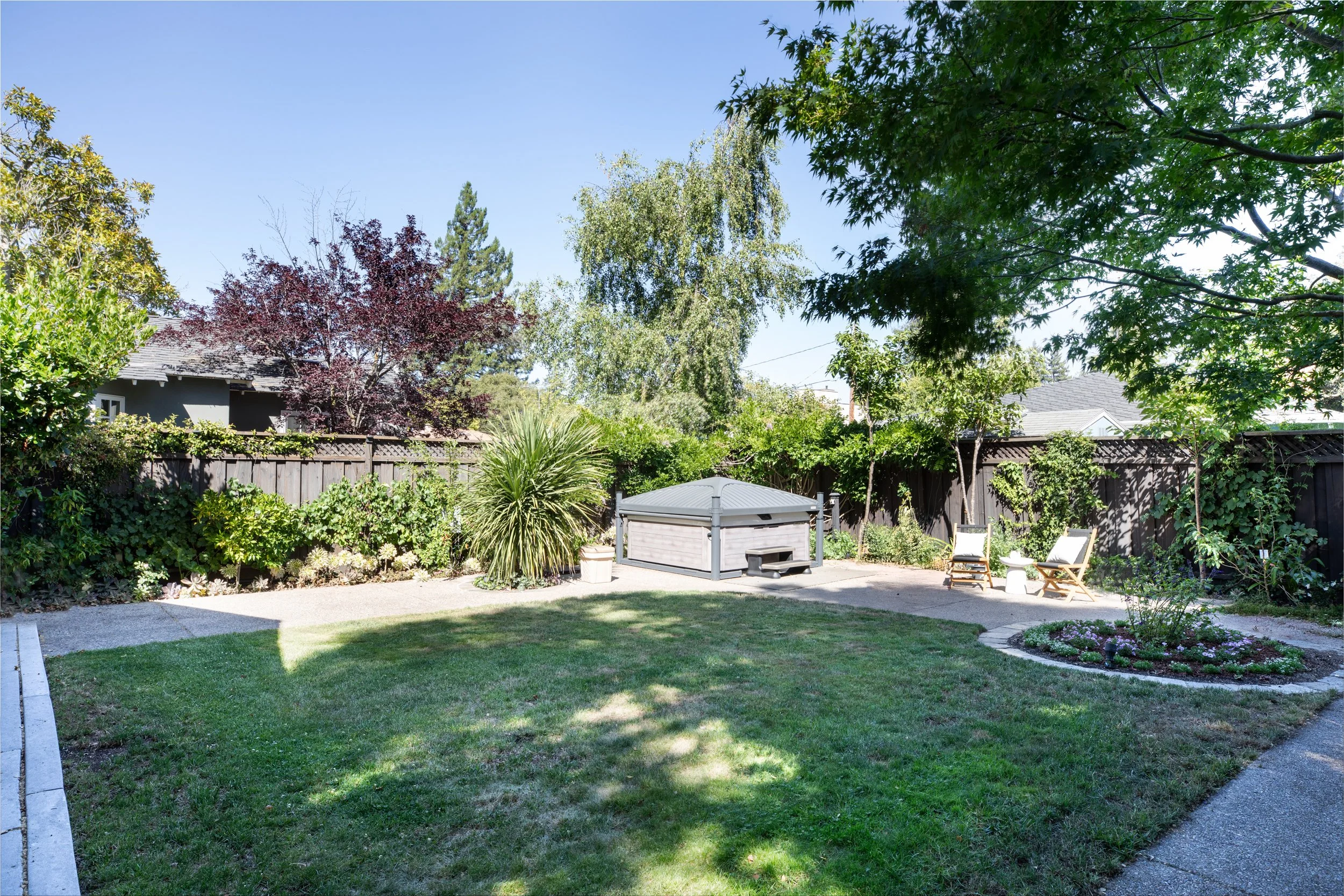 A backyard garden with a lush green lawn, surrounded by trees and bushes. There is a gazebo, garden chairs, and a small flower bed with purple flowers.