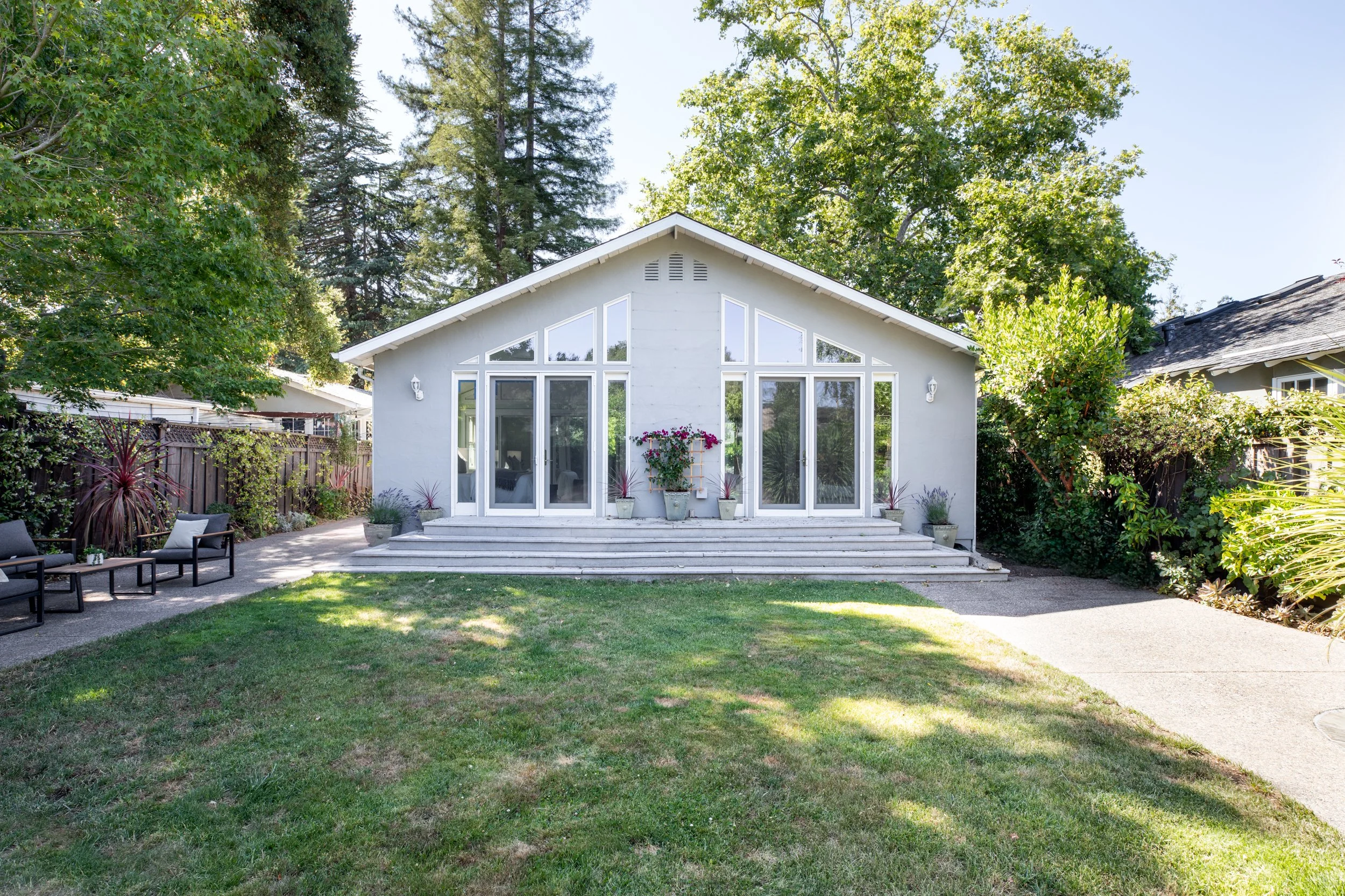 A white house with large windows and steps leading to a patio, surrounded by lush greenery and tall trees.