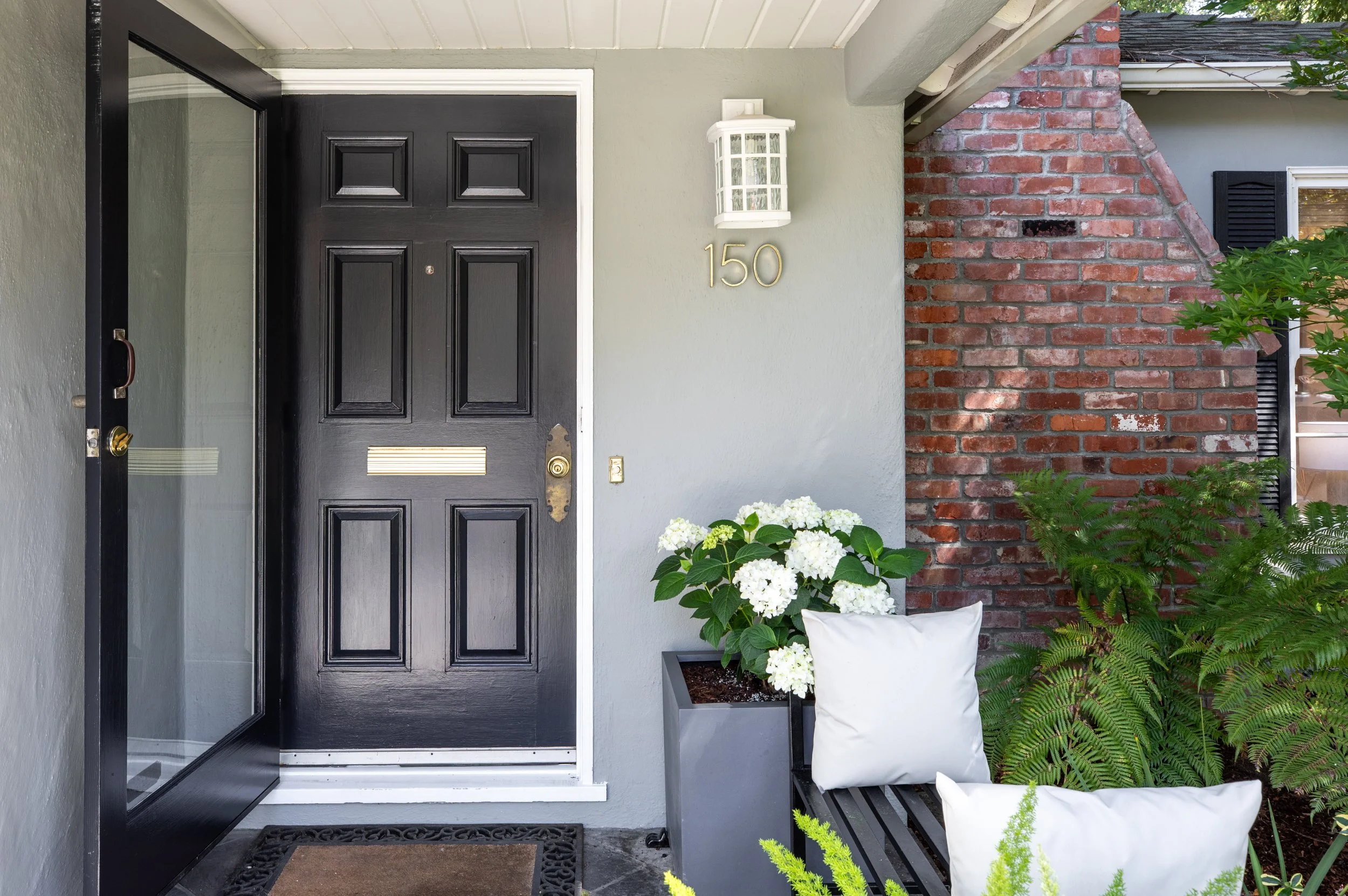 Front door of a house with black color, next to a wall with brick design, and lush green plants and white flowers in pots, with house number 150 displayed and a white porch light above.