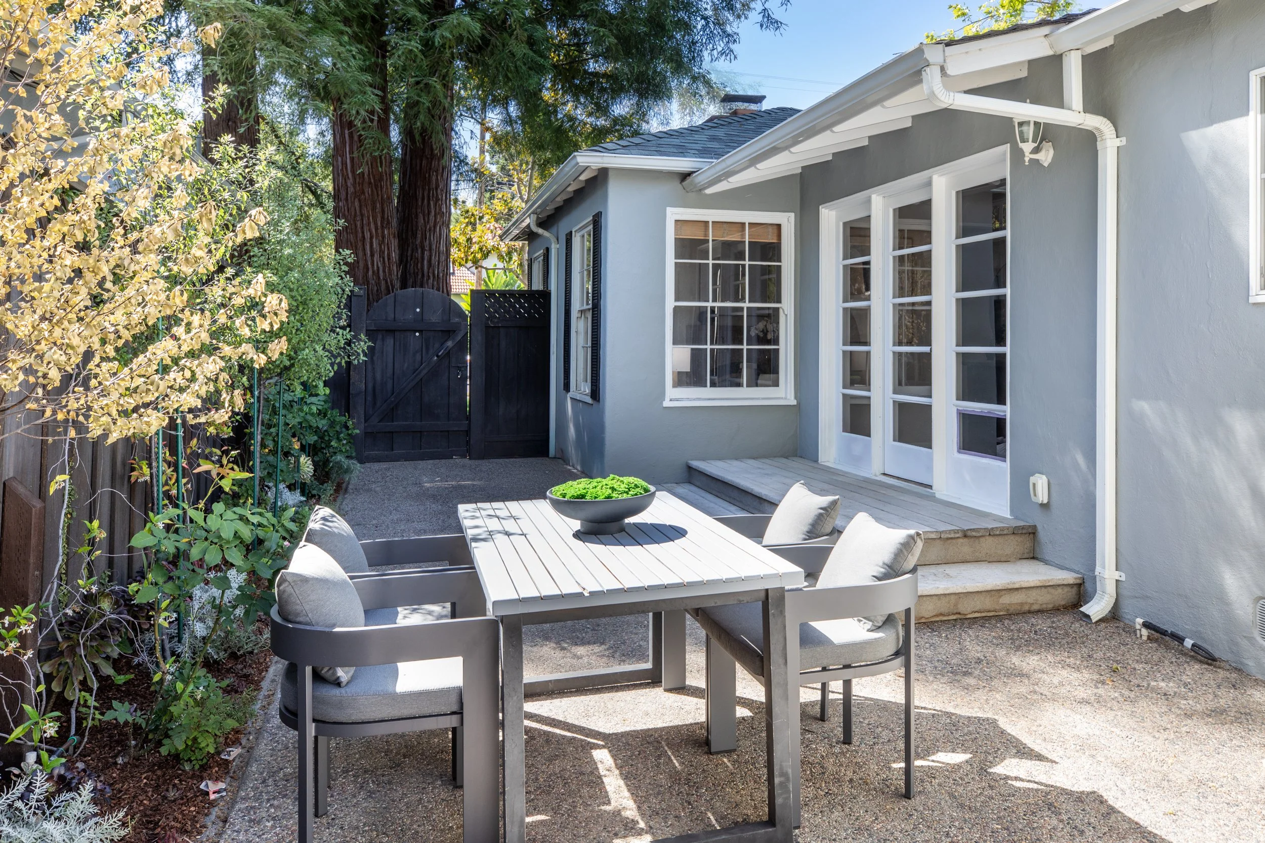A backyard patio with a white table and four gray chairs, surrounded by trees and plants, with a gray house featuring large windows and a black fence with a wooden gate.