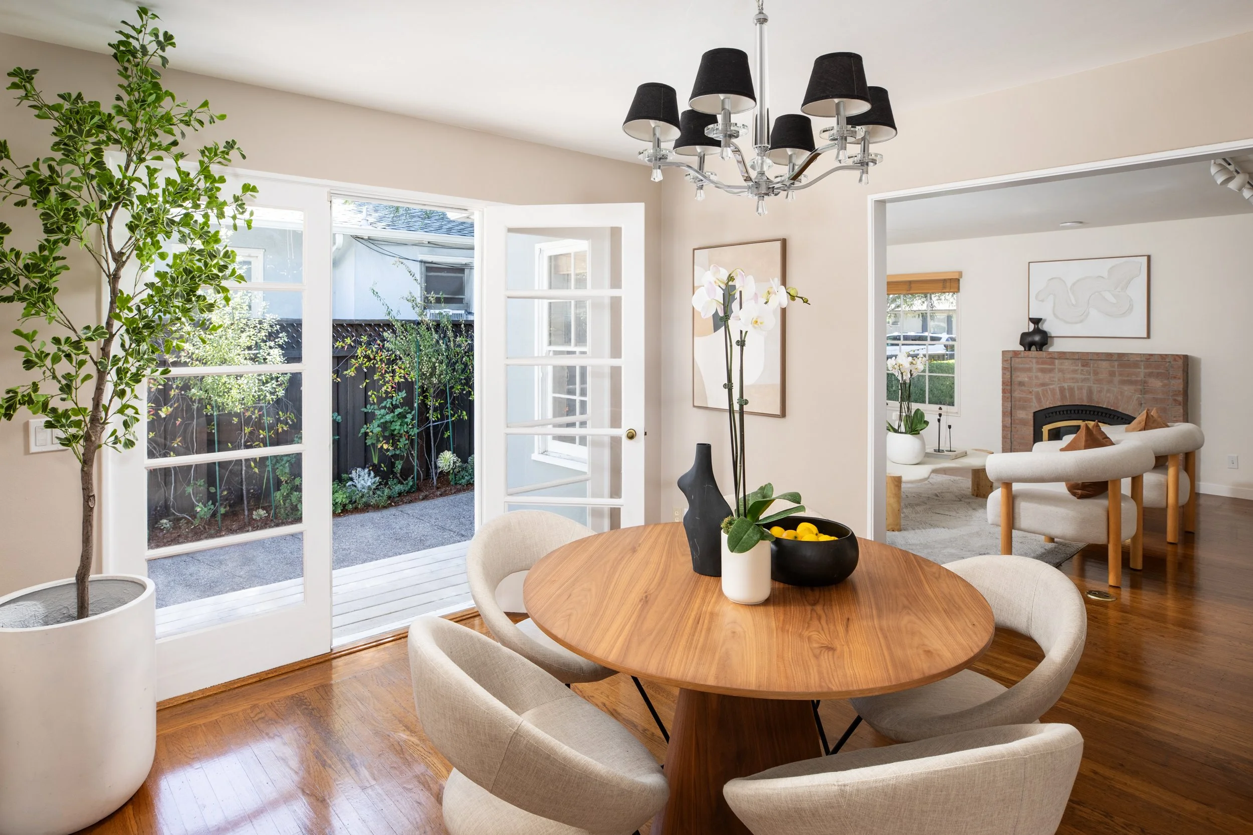A dining area with a round wooden table surrounded by beige upholstered chairs, a potted plant, and a black vase with white orchids on the table. French doors open to a small garden with greenery. In the background, a living room with a brick firepla
