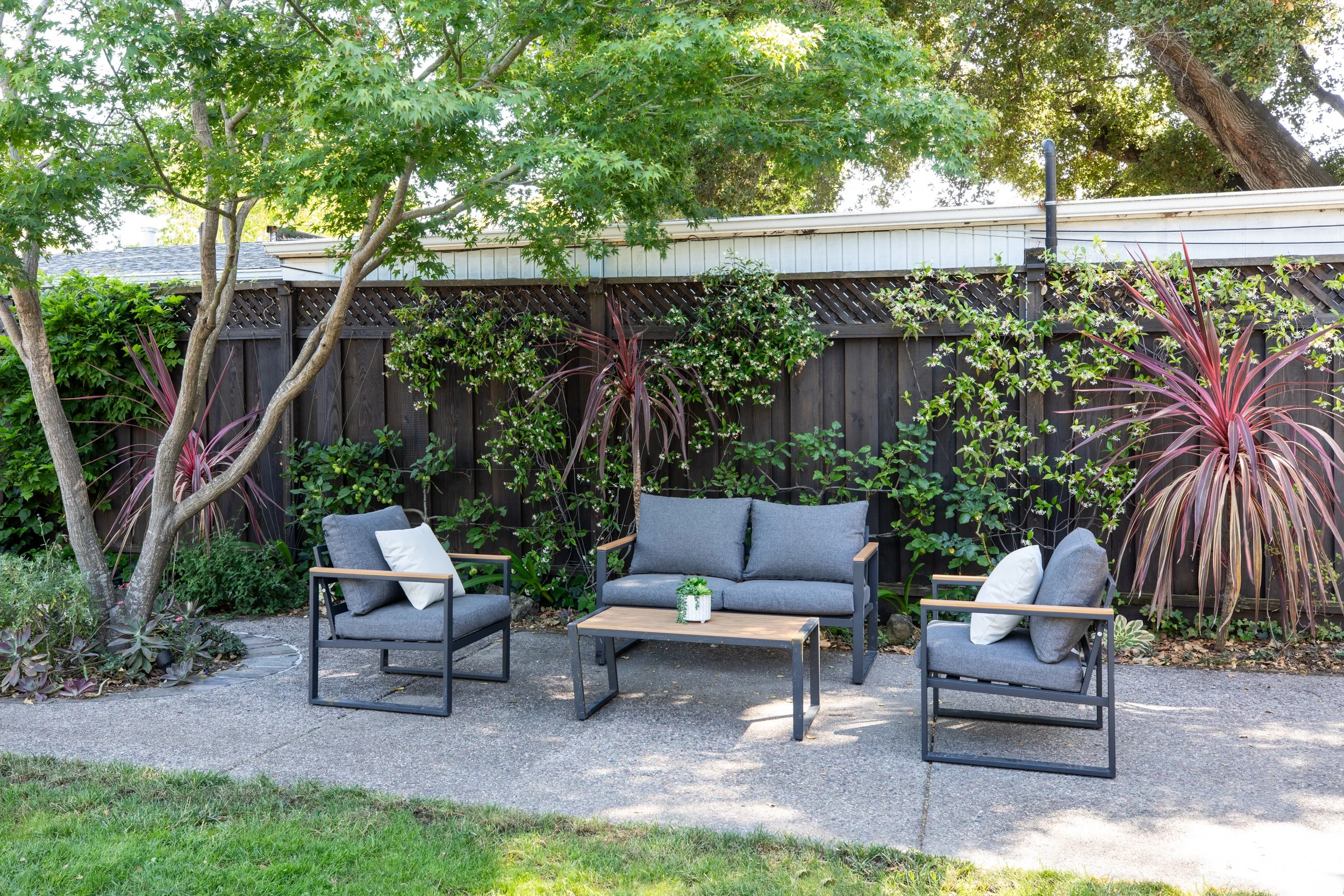 A backyard patio with grey outdoor furniture including a loveseat and two chairs, a wooden coffee table, surrounded by green trees and plants, with a wooden fence and a house with white siding in the background.