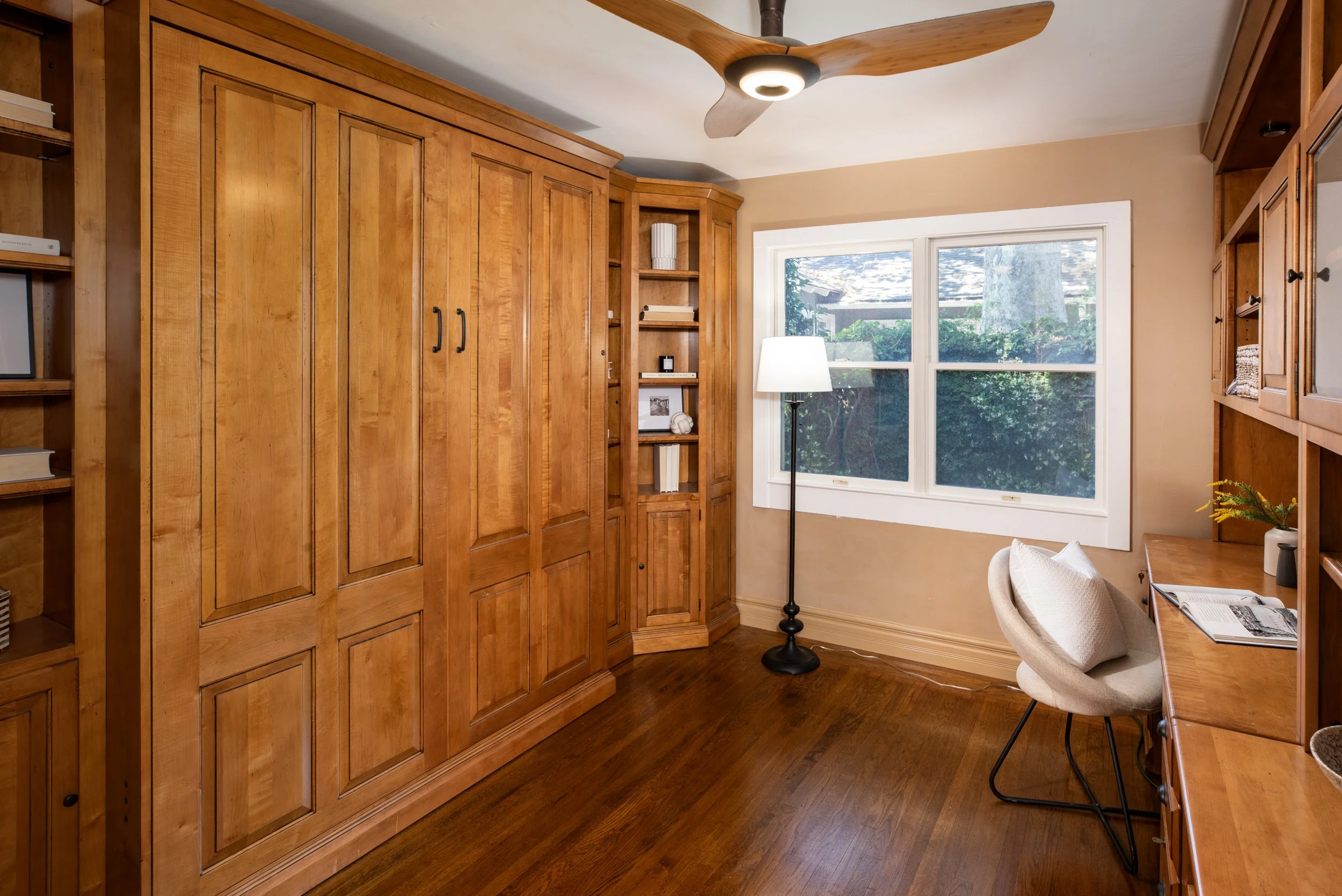Room with built-in wooden cabinets, a large window, a floor lamp, a cozy armchair, and a wooden desk.