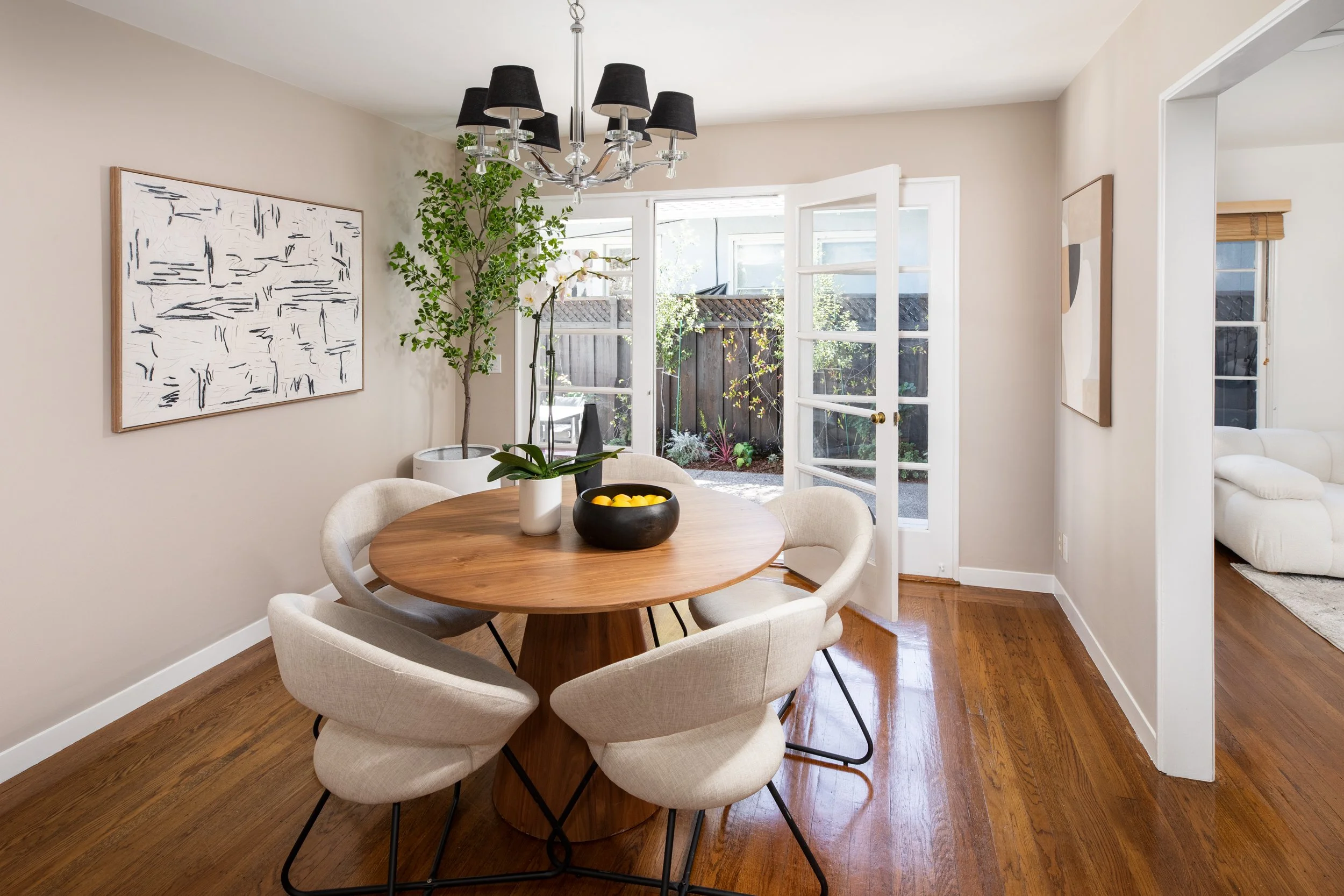 Dining room with round wooden table surrounded by beige upholstered chairs, large window with open glass door leading outside, artwork on the wall, indoor plant, chandelier, hardwood floors.