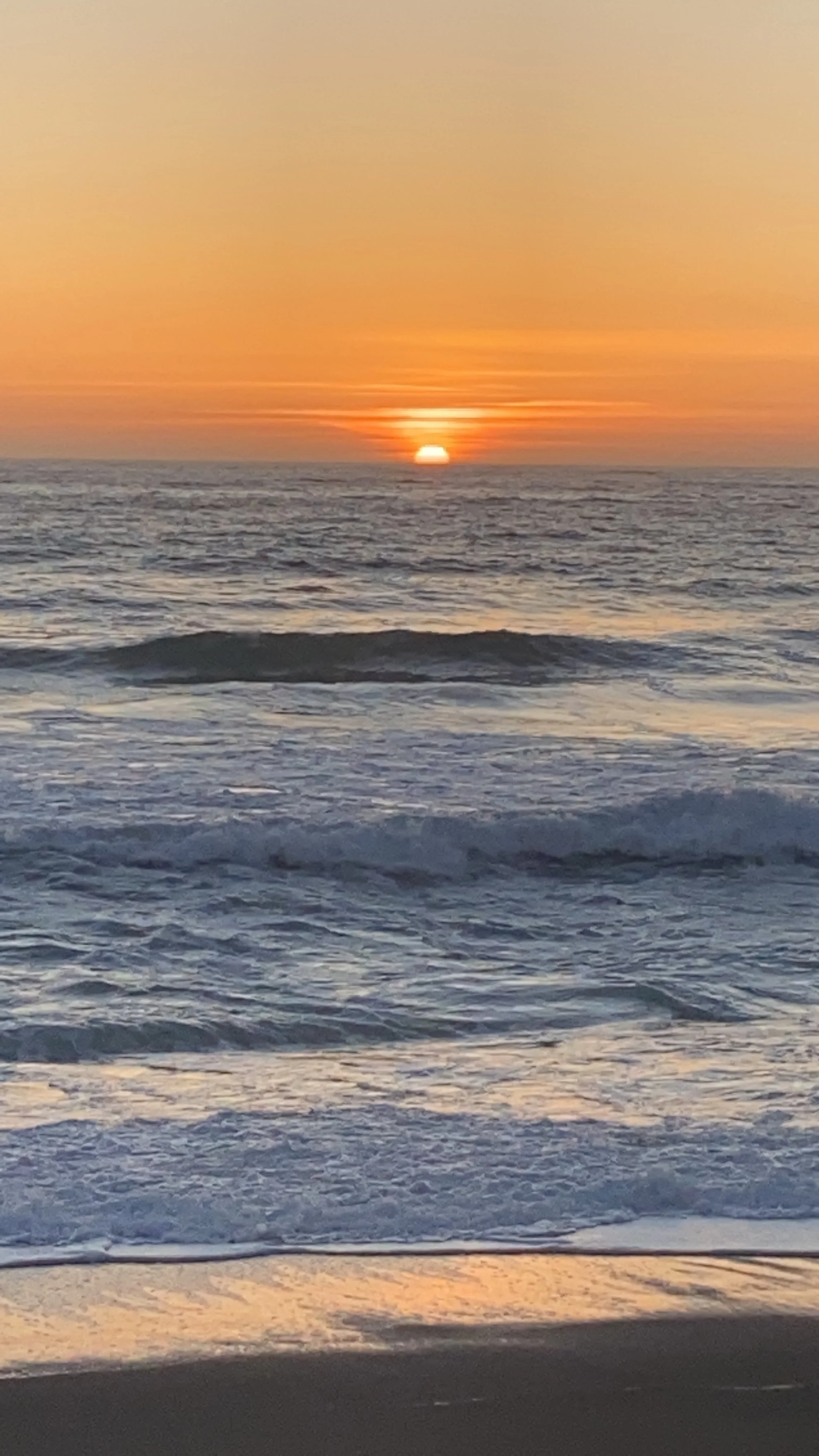 A sunset over the ocean with gentle waves in the foreground and the sun partially visible on the horizon, casting a warm orange glow across the sky and water.