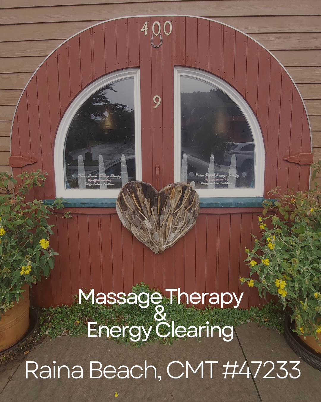The storefront of Raina Beach Massage Therapy  in Pacifica CA , it's a red arched door featuring two windows, a wooden heart decoration, and potted plants with yellow flowers outside. The window says by appointment only.