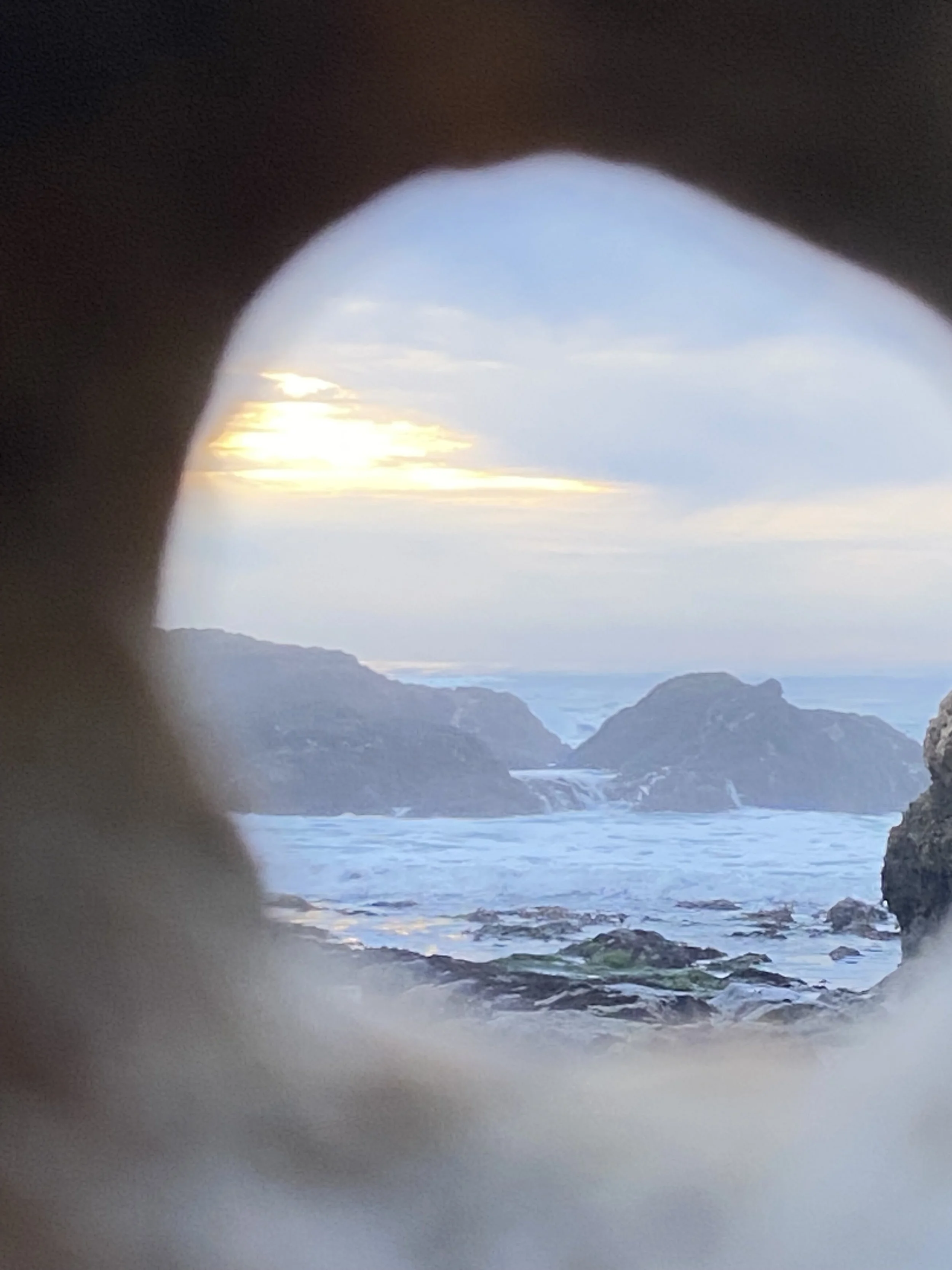 View through a small, circular opening showing a coastline with rocks, water, and distant hills under a cloudy sky during sunset.