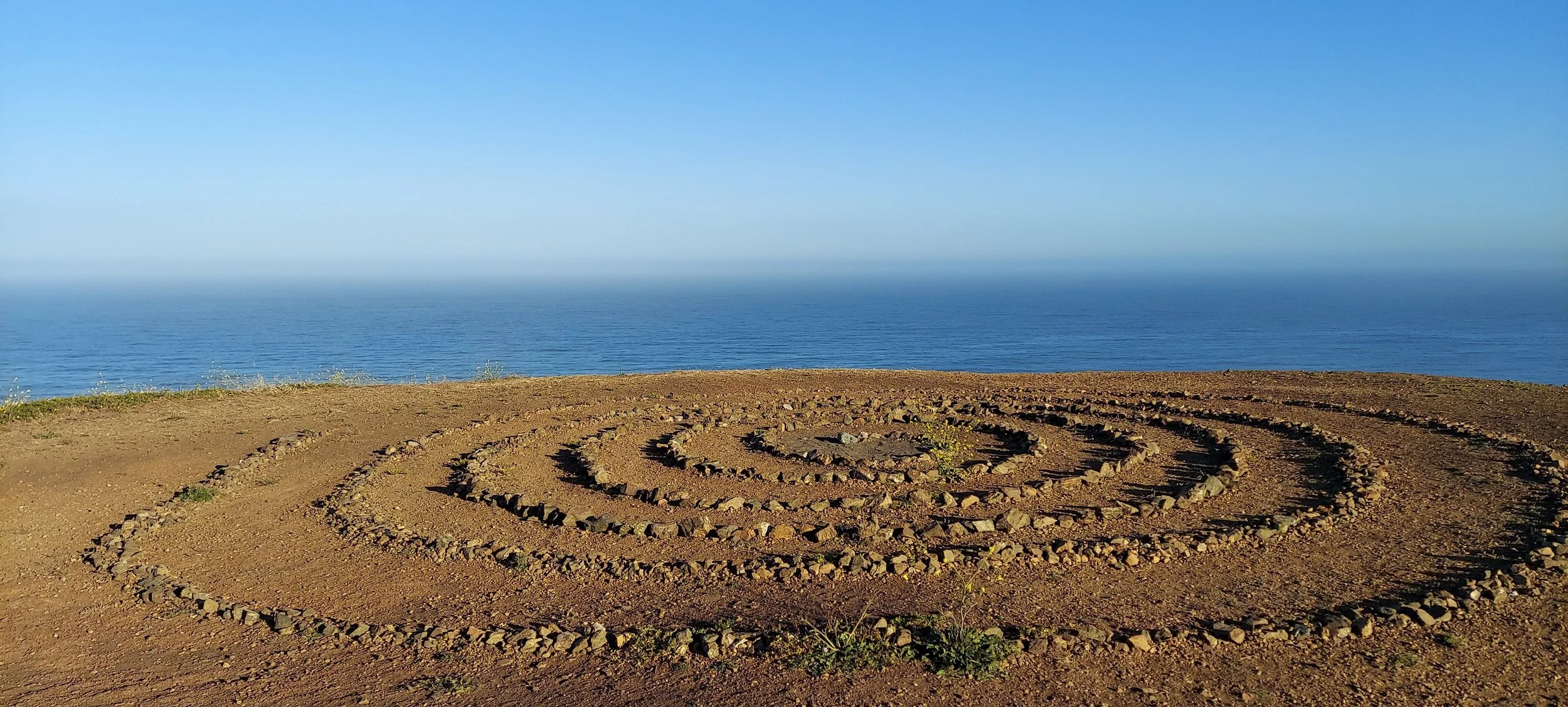 A spiral-shaped rock formation on a dirt ground overlooking a calm ocean with a clear blue sky.