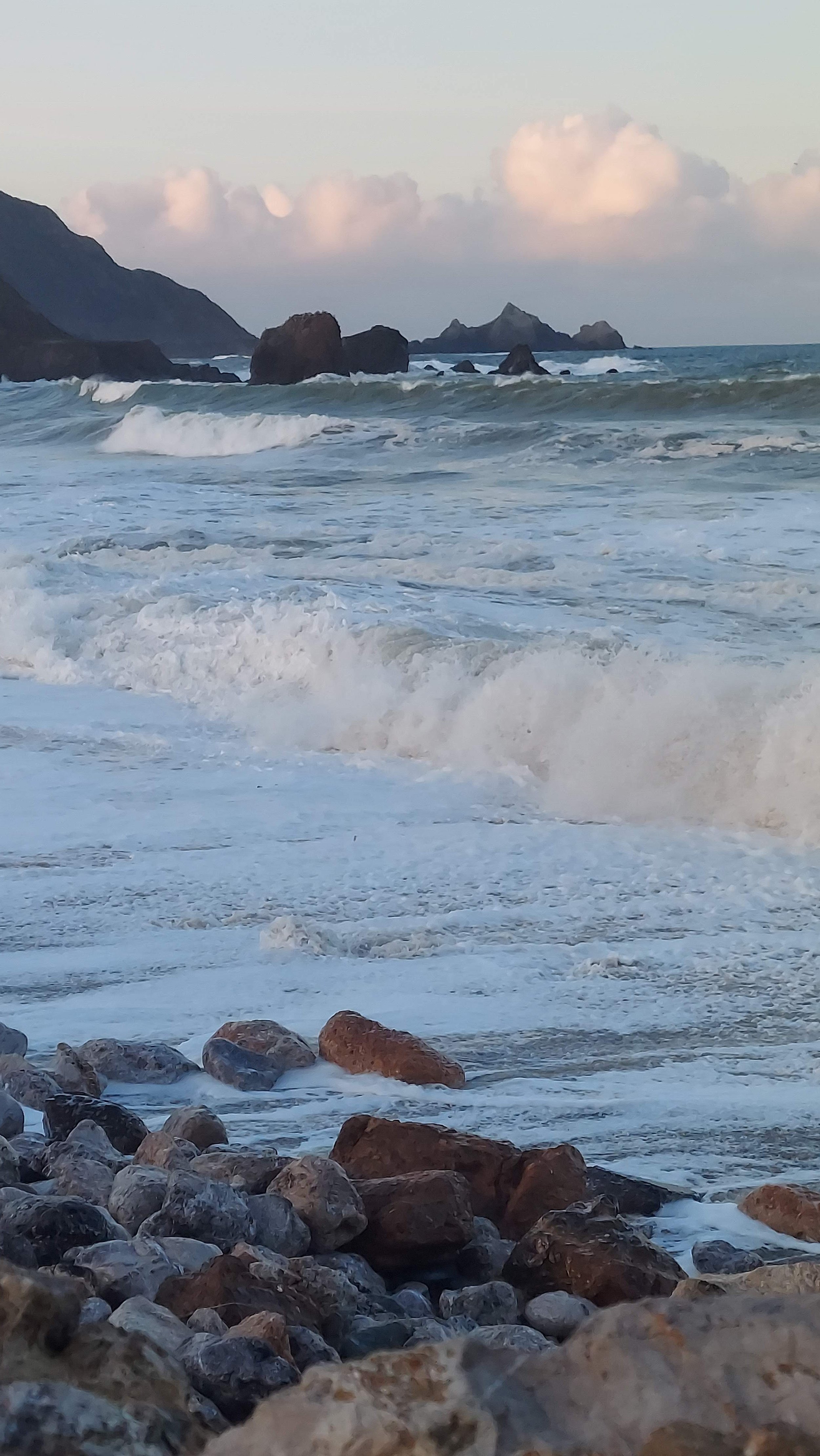 Rocky shoreline with waves crashing against the rocks, ocean and distant mountains with clouds in the sky.