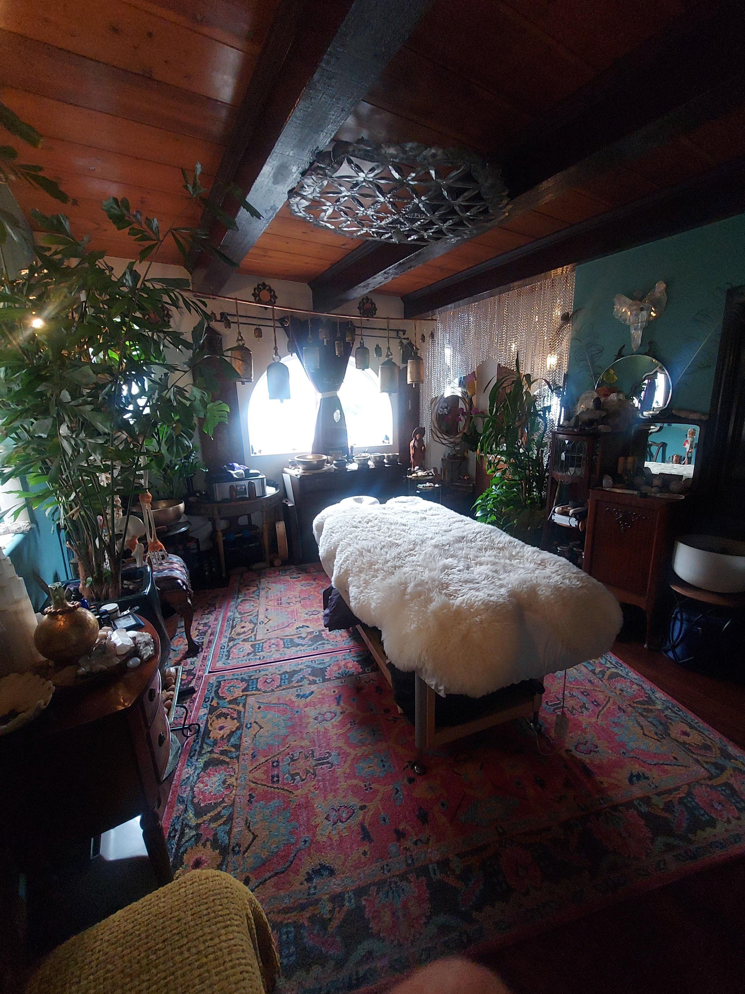 A cozy room with a wooden ceiling, large window with curtains, a massage table with a fluffy cover, a colorful oriental rug, and various plants and decorative items.