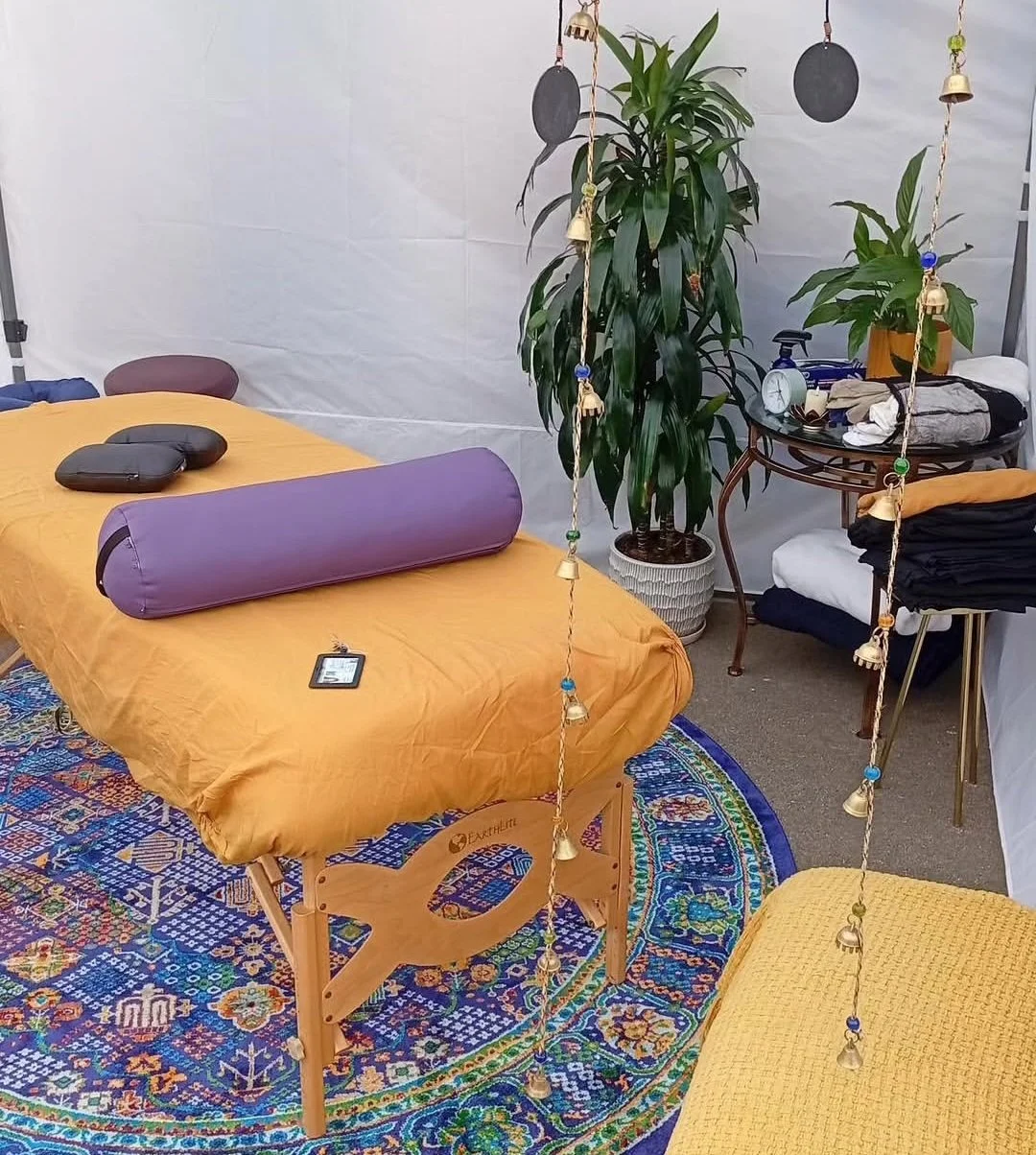 A massage table covered with a yellow sheet, placed on a colorful round rug. There is a purple bolster on the table. To the right, a large potted plant, a small side table with folded clothes, a clock, and a bottle. Behind the table, decorative hanging ornaments are suspended from the ceiling.