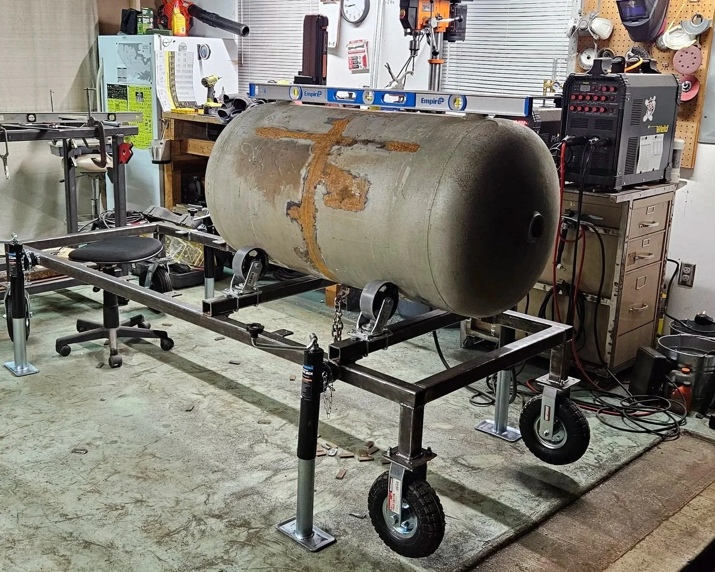 A large rusted tank on a wheeled metal frame in a workshop, with tools, a level, and a chair around it.