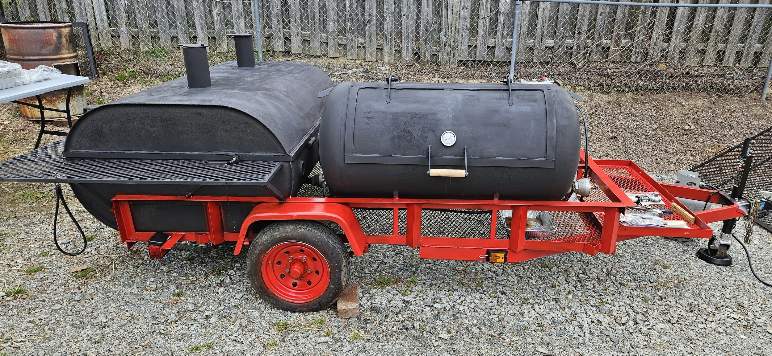 A mobile barbecue smoker on a red trailer with two large black smoking chambers, a firebox, and a temperature gauge, set up outdoors on gravel ground.