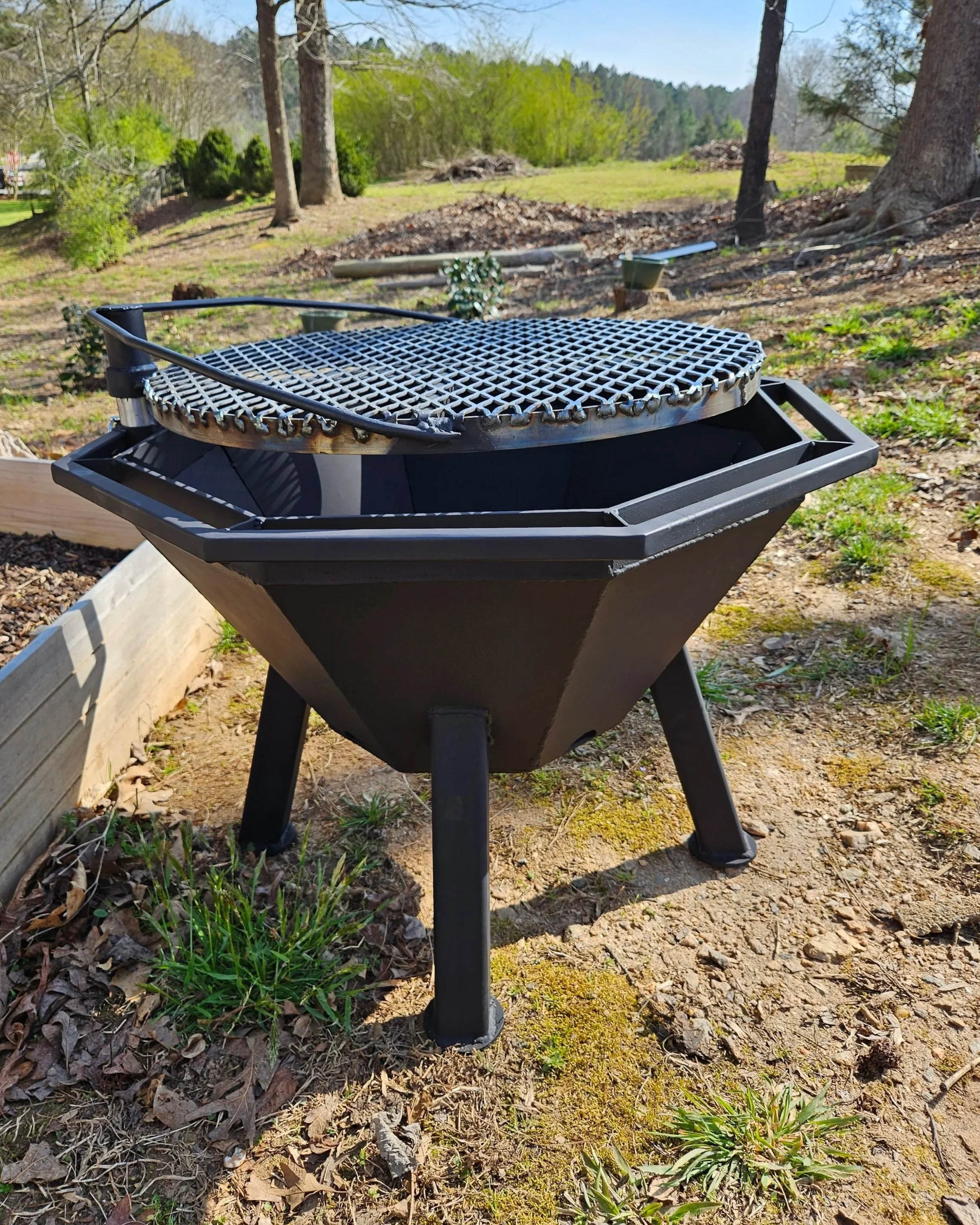 An outdoor barbecue grill with a metal cooking surface stands on a patch of dirt and grass, surrounded by trees and hills in the background.