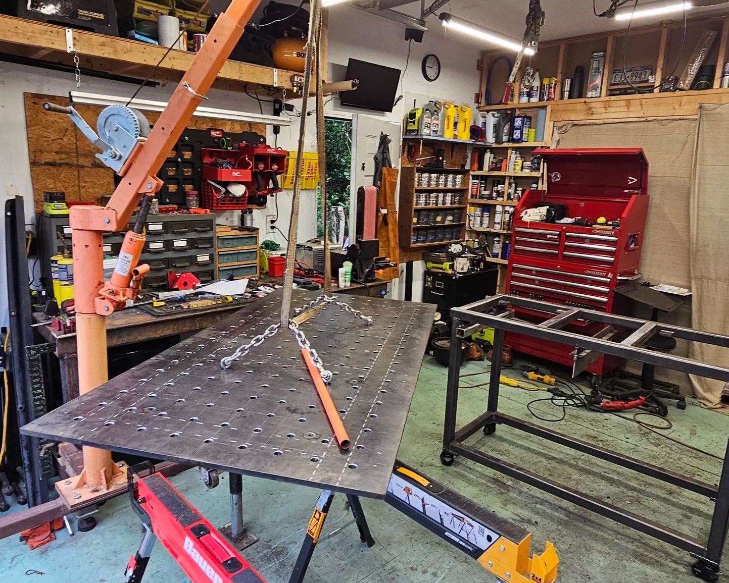 A workshop with a large metal table, a hoist with chains, and various tools and storage units. The shop is organized, with shelves filled with supplies and containers, and a red tool chest.