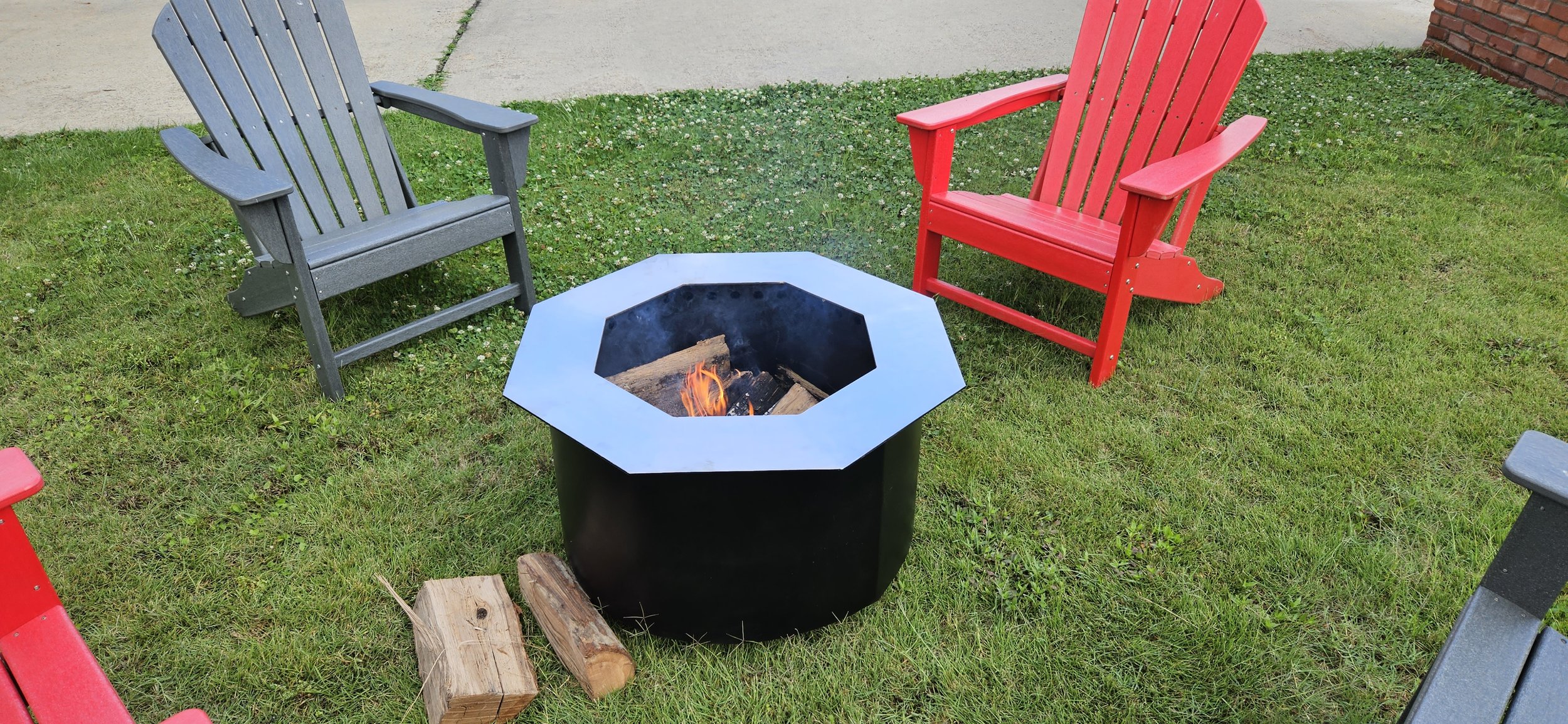 Two Adirondack chairs, one red and one gray, are placed on grass near a fire pit with a small fire burning inside. There are a couple of pieces of firewood on the ground nearby.