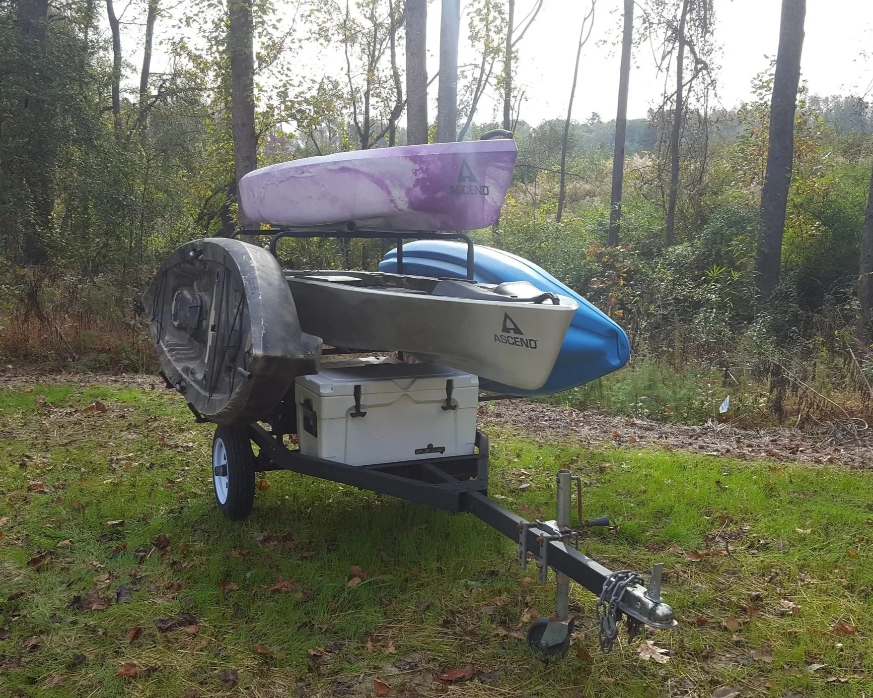 A small trailer with three stacked kayaks, a cooler, and other gear, parked on a grassy area with trees in the background.