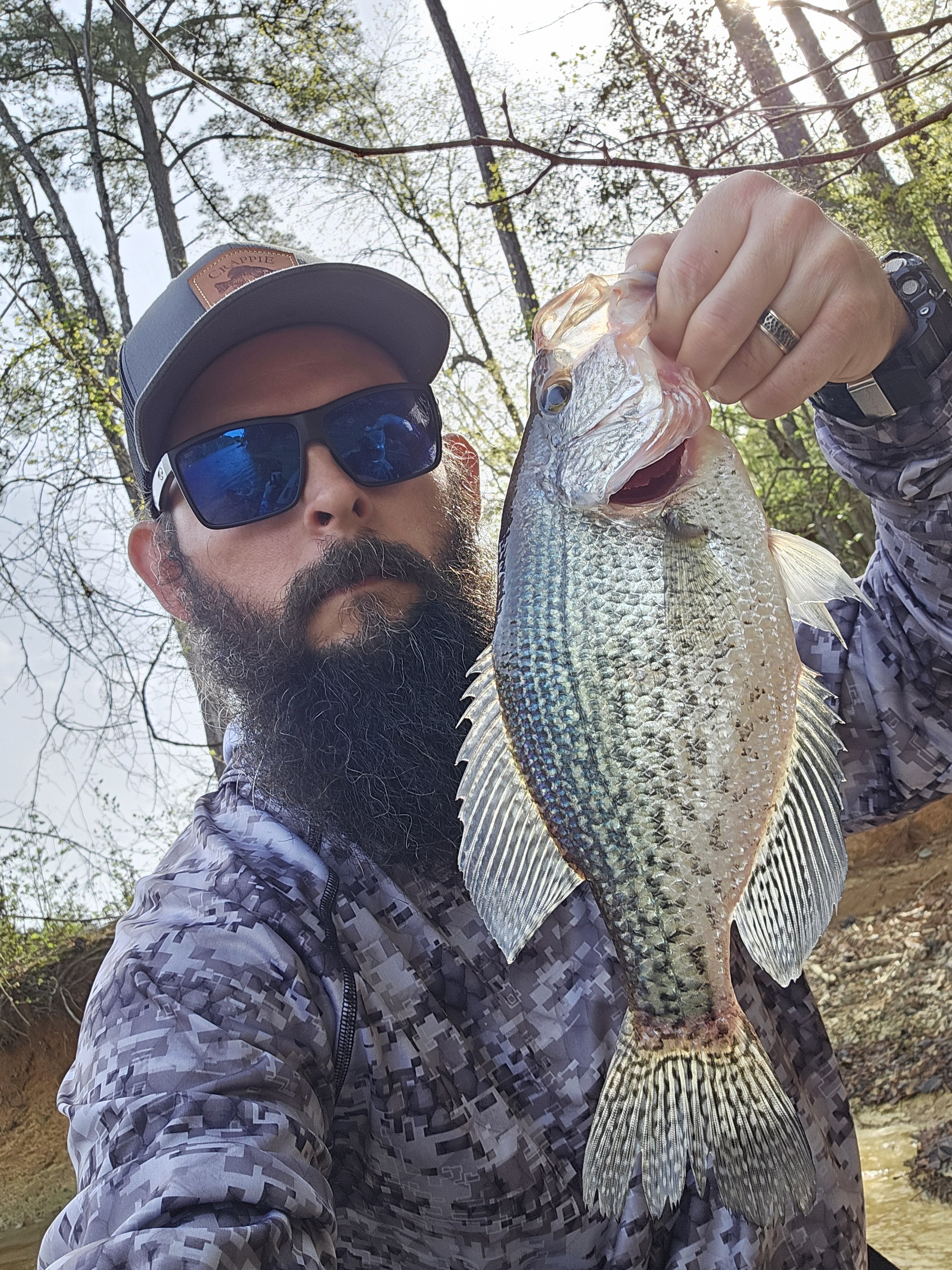 A man with a beard wearing sunglasses and a cap holding a large fish, outdoors with trees and a river in the background.
