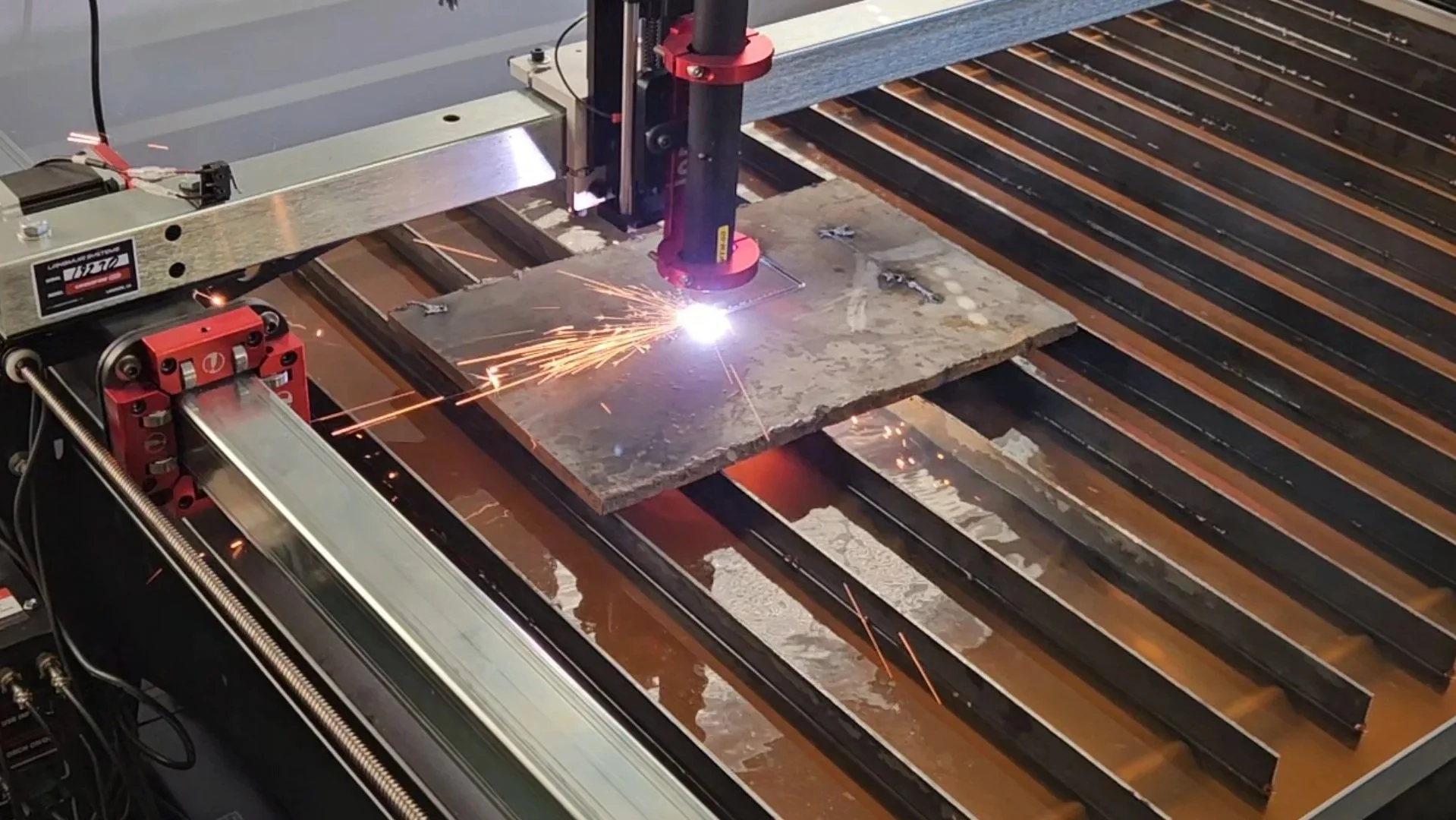 A CNC plasma cutting machine cuts through a metal sheet, producing sparks and bright light at the cutting point, with orange and black striped materials underneath.