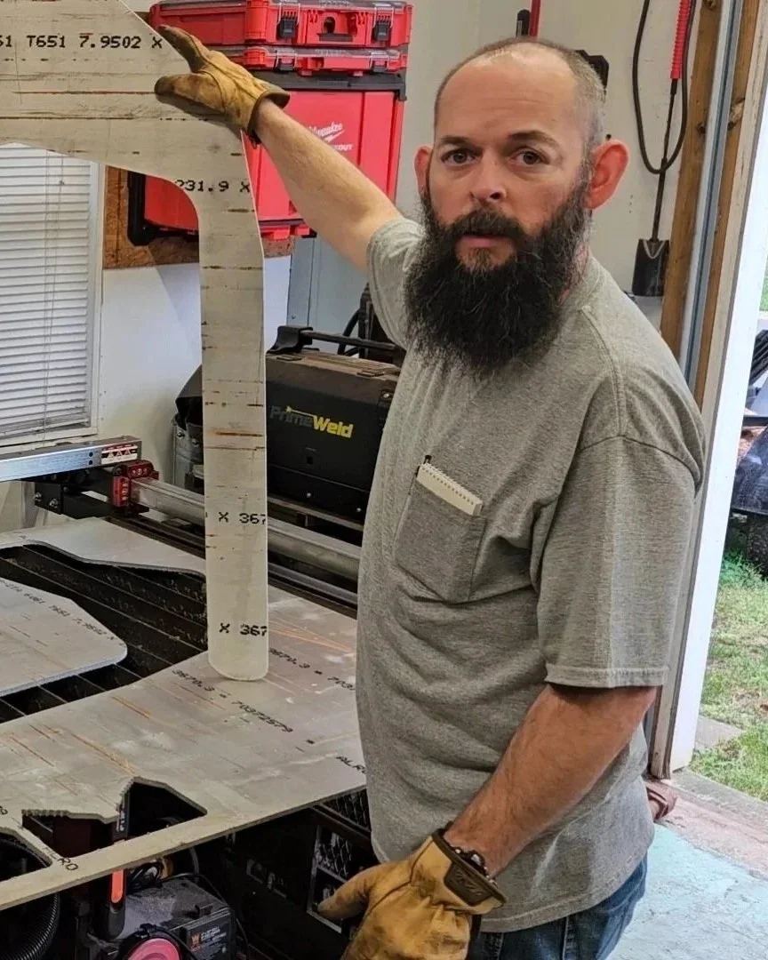 A man with a beard wearing a gray t-shirt and yellow work gloves, holding a long piece of wood in a workshop with tools and equipment visible behind him.