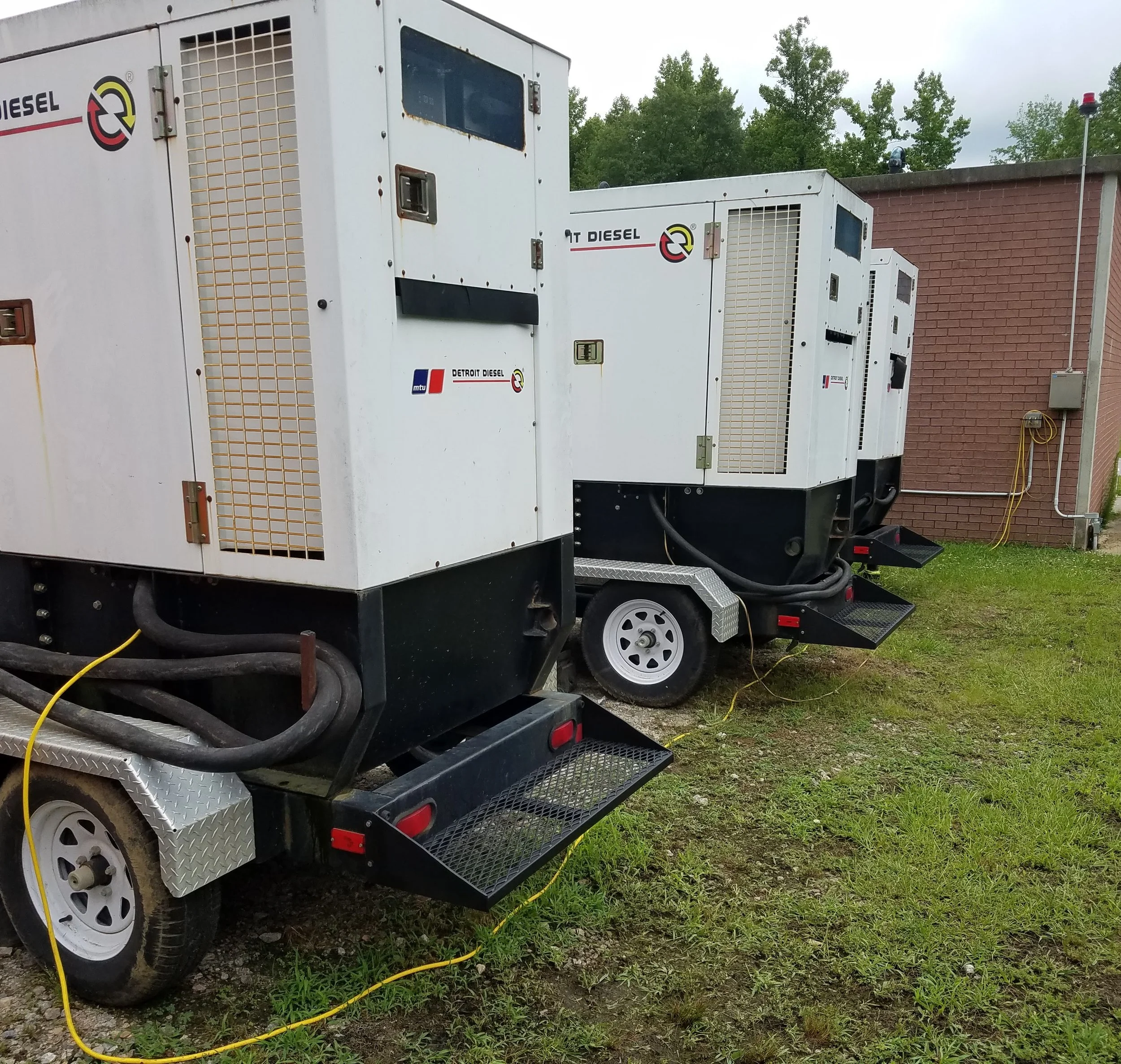 Two large white diesel generator units on a trailer parked on grass near a brick building, with trees in the background.