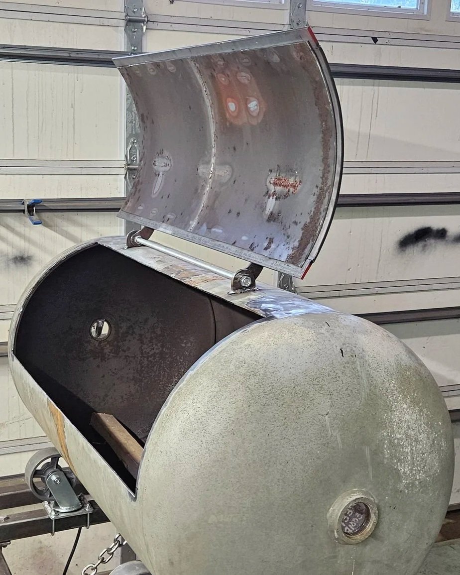 A cylindrical metal smoker with a hinged lid open, situated in a garage with a white, paneled garage door in the background.