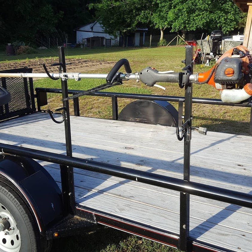 A flatbed utility trailer with a weed trimmer mounted on the front rail, parked on grass with trees and a building in the background.