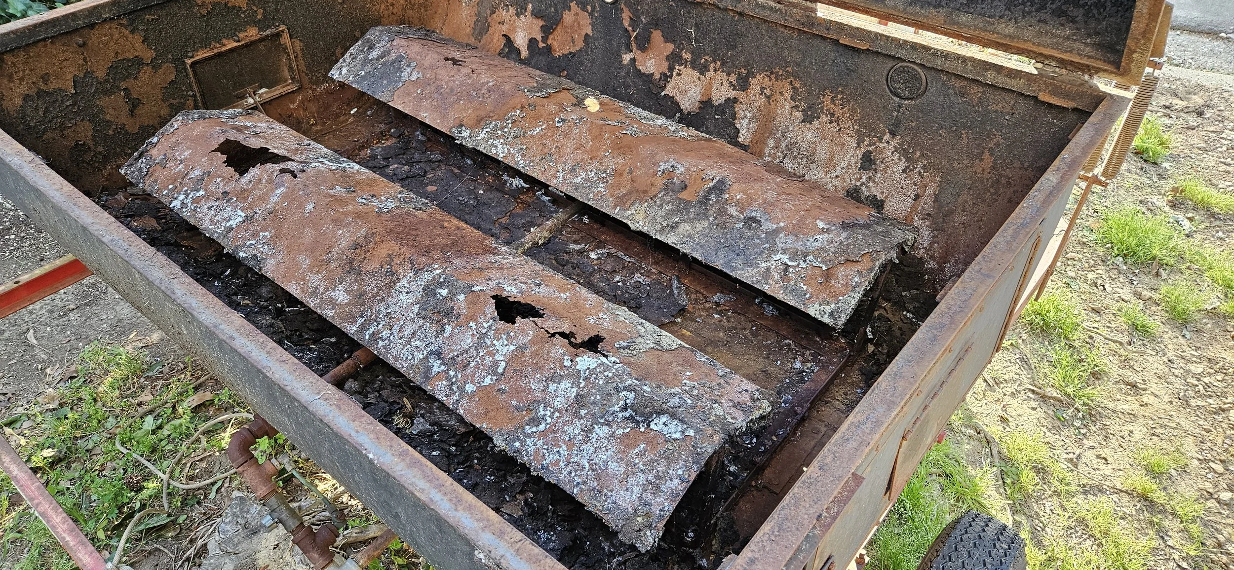 A rusty, damaged metal barbecue grill with peeling paint and corroded grates, sitting outdoors on grass and dirt.