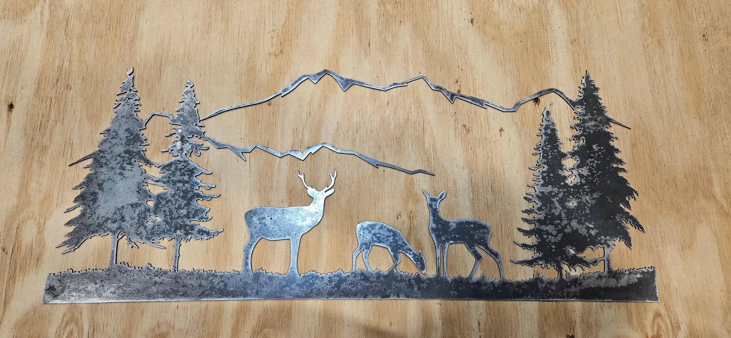Metal wall art depicting trees, mountains, and three deer on a wooden background.