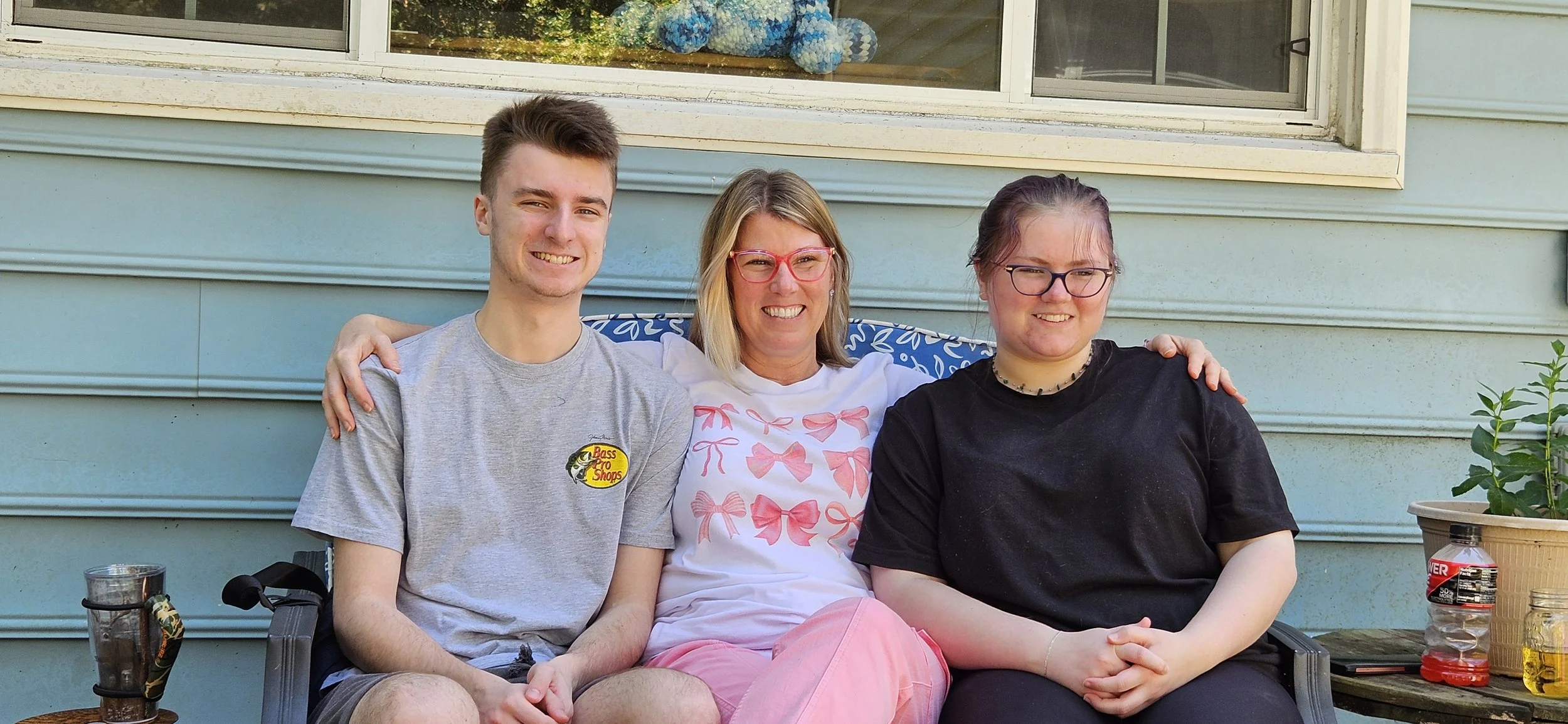 Three people sitting on a porch outside a house. A young man on the left wearing a gray t-shirt with a Bass Pro Shops logo, a woman in the middle with blonde hair and red glasses wearing a white shirt with pink bows, and a young woman on the right with glasses and dark hair wearing a black shirt. They are smiling and have their arms around each other.