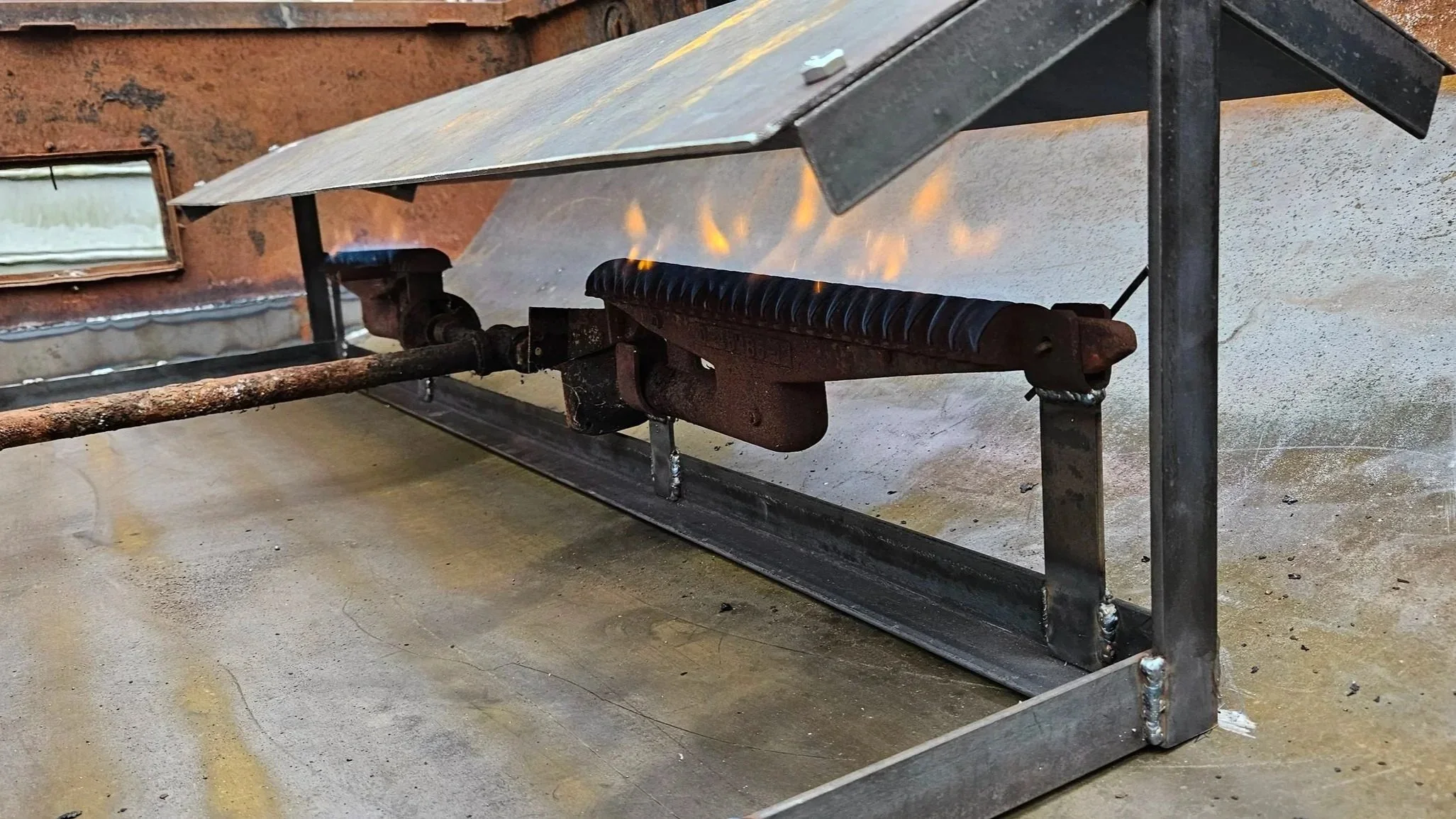 A metal forge with a small open flame underneath, situated on a concrete floor in a workshop or industrial setting.