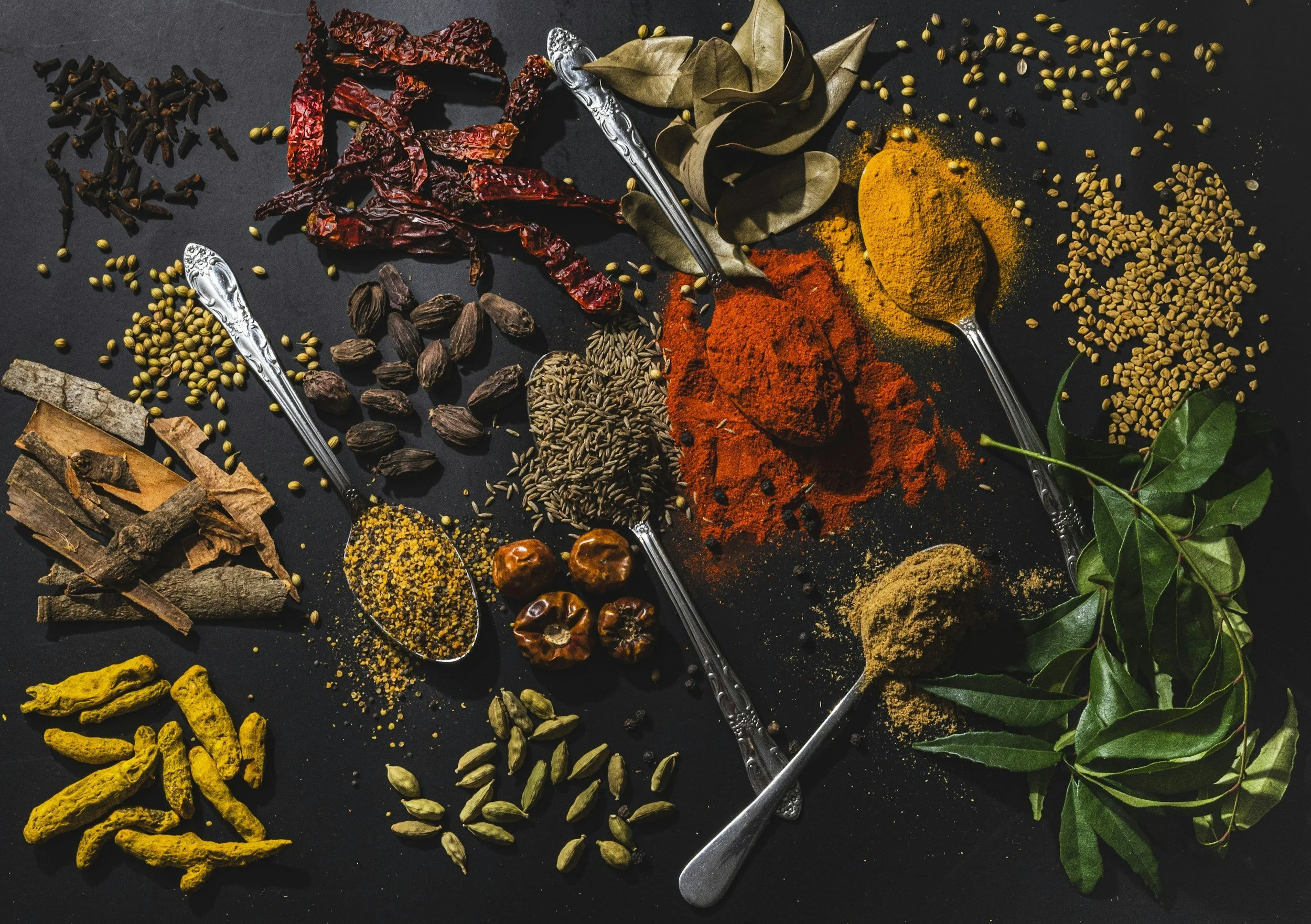 Assorted spices and herbs arranged on a dark surface, including dried red chili peppers, turmeric, curry powder, cumin seeds, coriander seeds, bay leaves, cinnamon sticks, cardamom pods, cloves, nutmeg, and dried turmeric roots, with vintage silver spoons holding some spices.