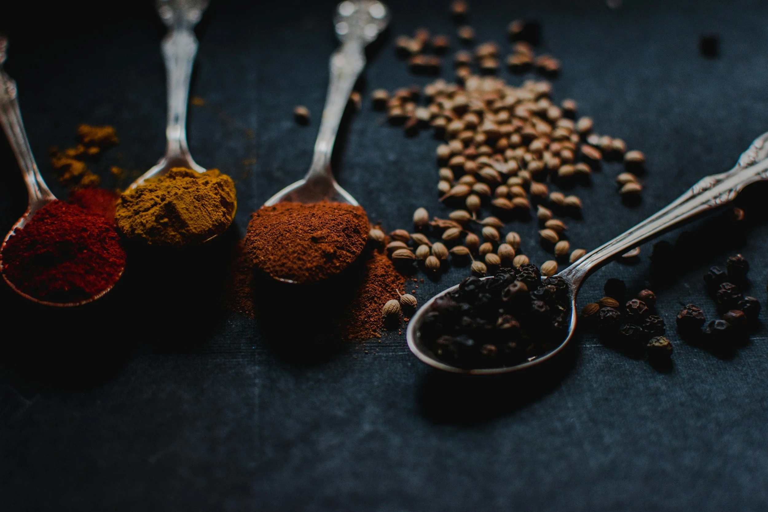 Spoons filled with ground spices, and scattered black peppercorns on a dark surface.