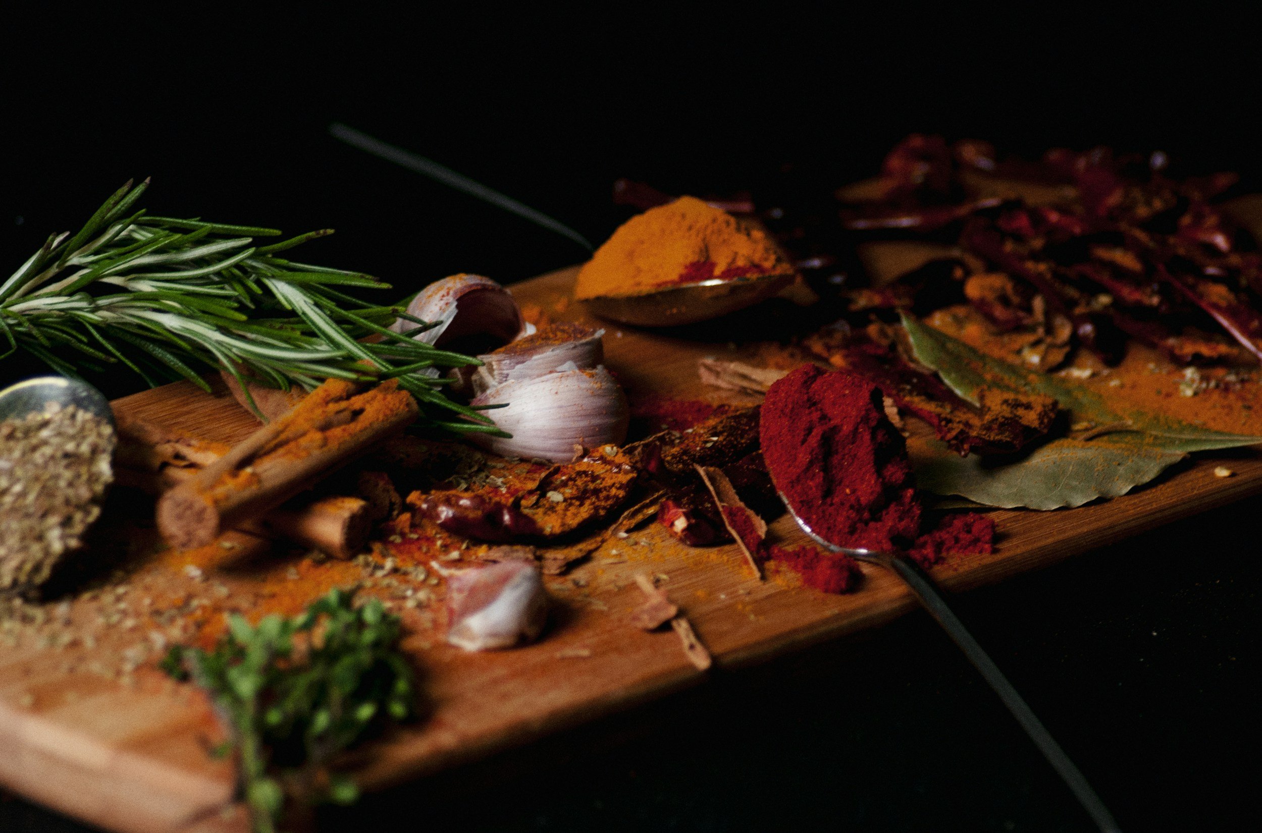 A wooden board with fresh herbs, garlic, spices, and dried chili peppers arranged on it against a black background.
