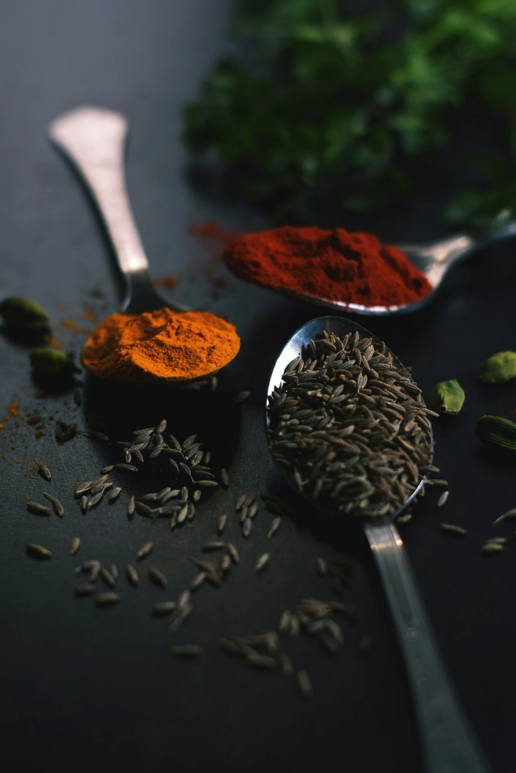 Spoons filled with spices, including red chili powder, turmeric powder, and cumin seeds, on a dark surface with scattered cumin seeds and some green herbs in the background.