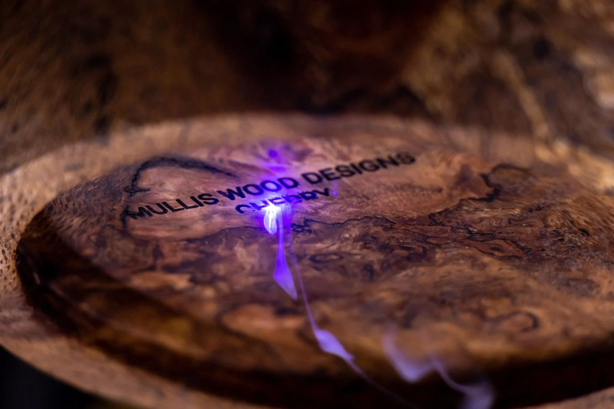 Close-up of a piece of wood with text labeled 'MULLIS WOOD DESIGNS' illuminated by a purple laser.