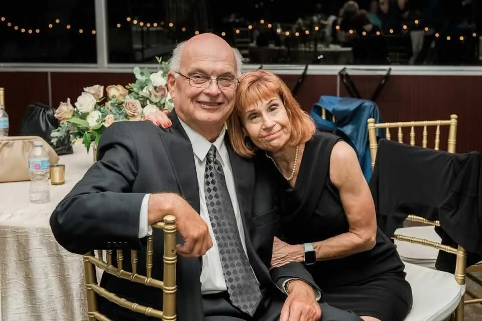 Naomi and her husband sit together at a formal event, smiling. He is in a dark suit with a patterned tie, and she is in a sleeveless black dress with a pearl necklace. They are seated at a table with a floral centerpiece and gold chairs.