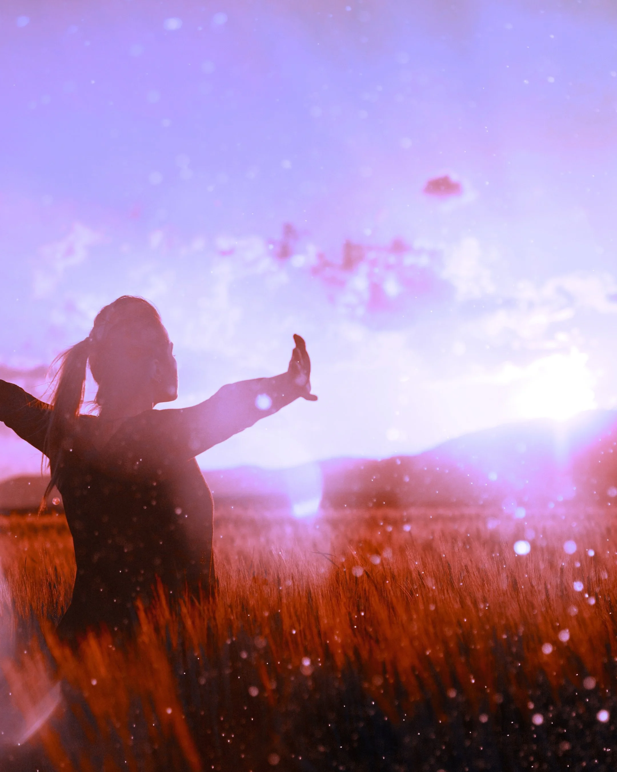 A person with long hair has their arms open at sunrise, with a field of tall grass in the foreground and a colorful sky with clouds and distant mountains in the background.
