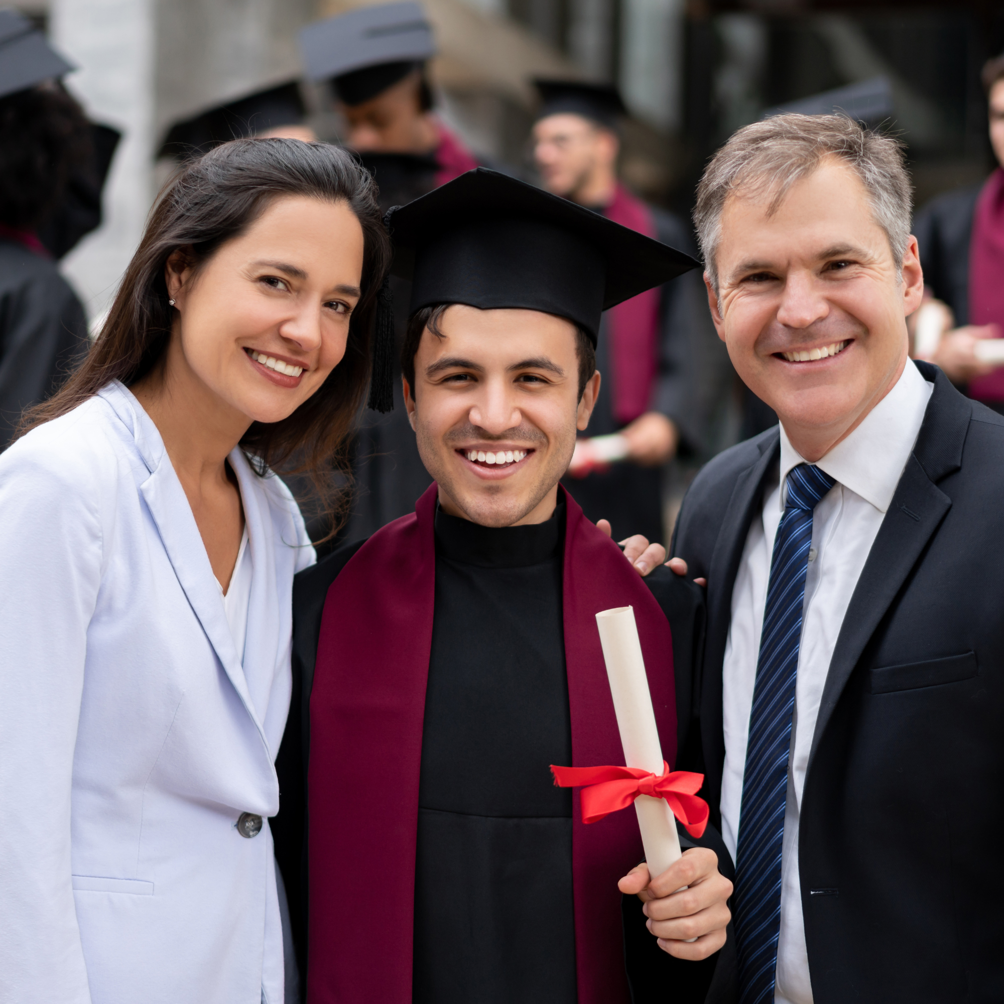 A group of people at a graduation ceremony, including a woman in a white suit, a man in a blue suit, and a graduate in a cap and gown, holding a diploma with a red ribbon.