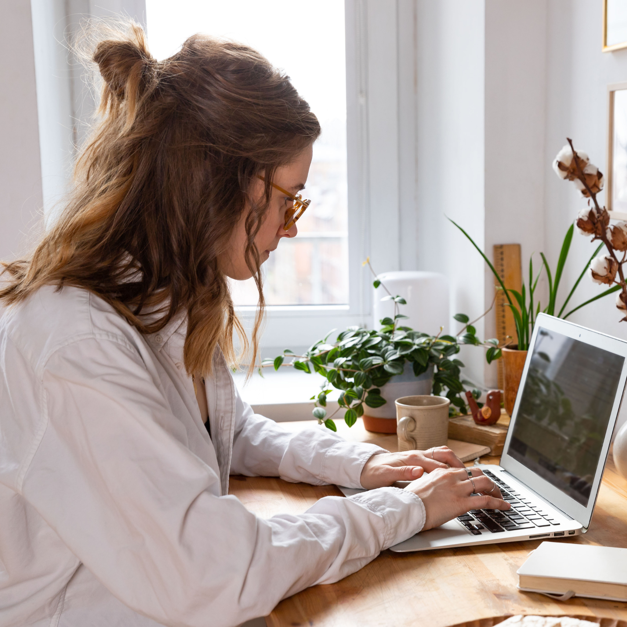 A woman with long, wavy, reddish-brown hair in a messy top knot wearing glasses, working on a laptop at a wooden desk near a window. The desk has potted plants, a book, and a tablet.