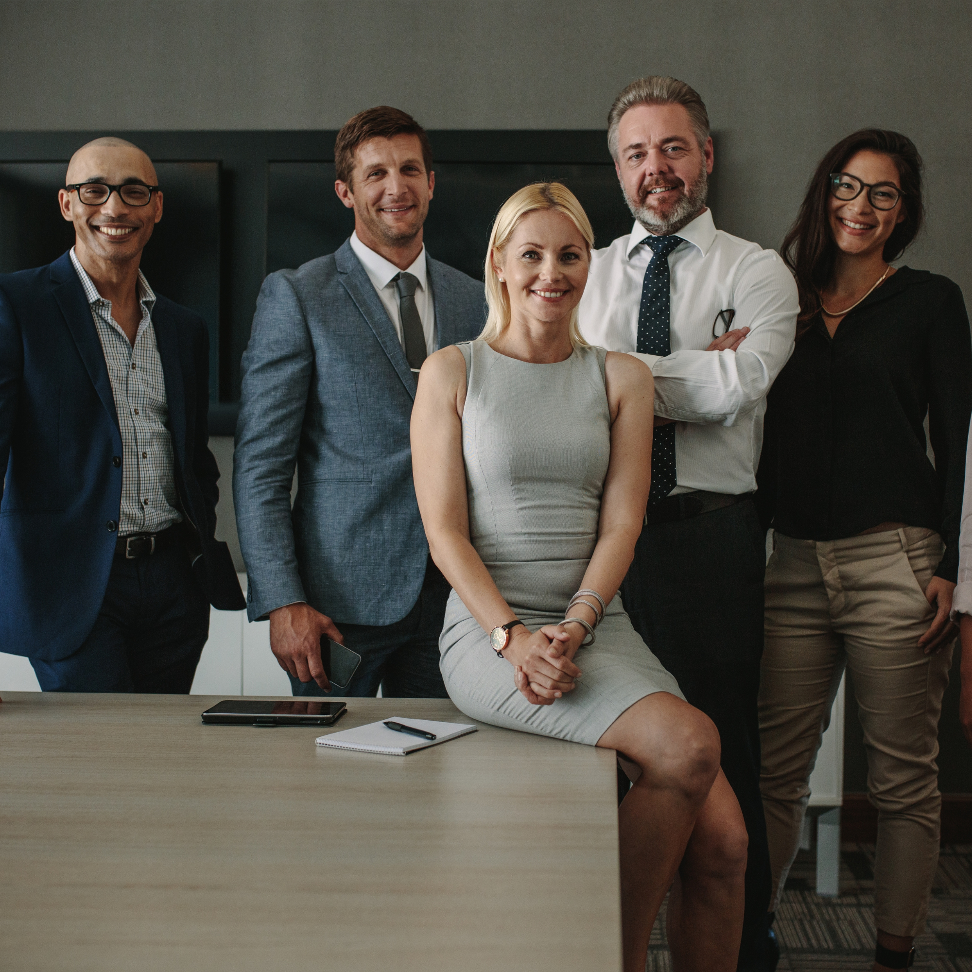 Group of five diverse business professionals, three men and two women, standing behind a woman sitting at a desk with a notepad, pen, tablet, and smartphone in an office setting.