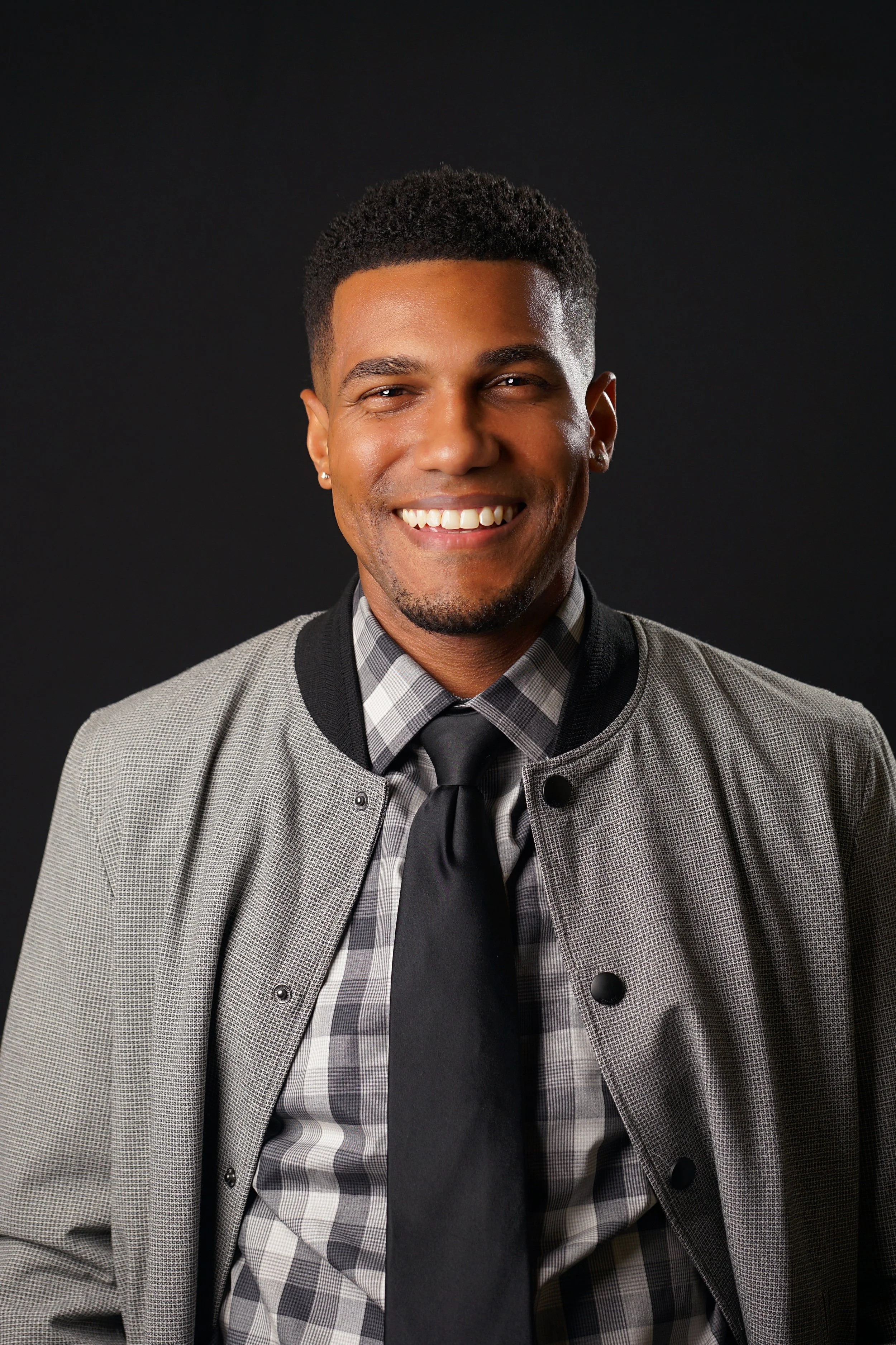 A smiling man with short curly black hair, wearing a gray checked blazer over a checkered shirt, black tie, and black buttons, set against a plain black background.