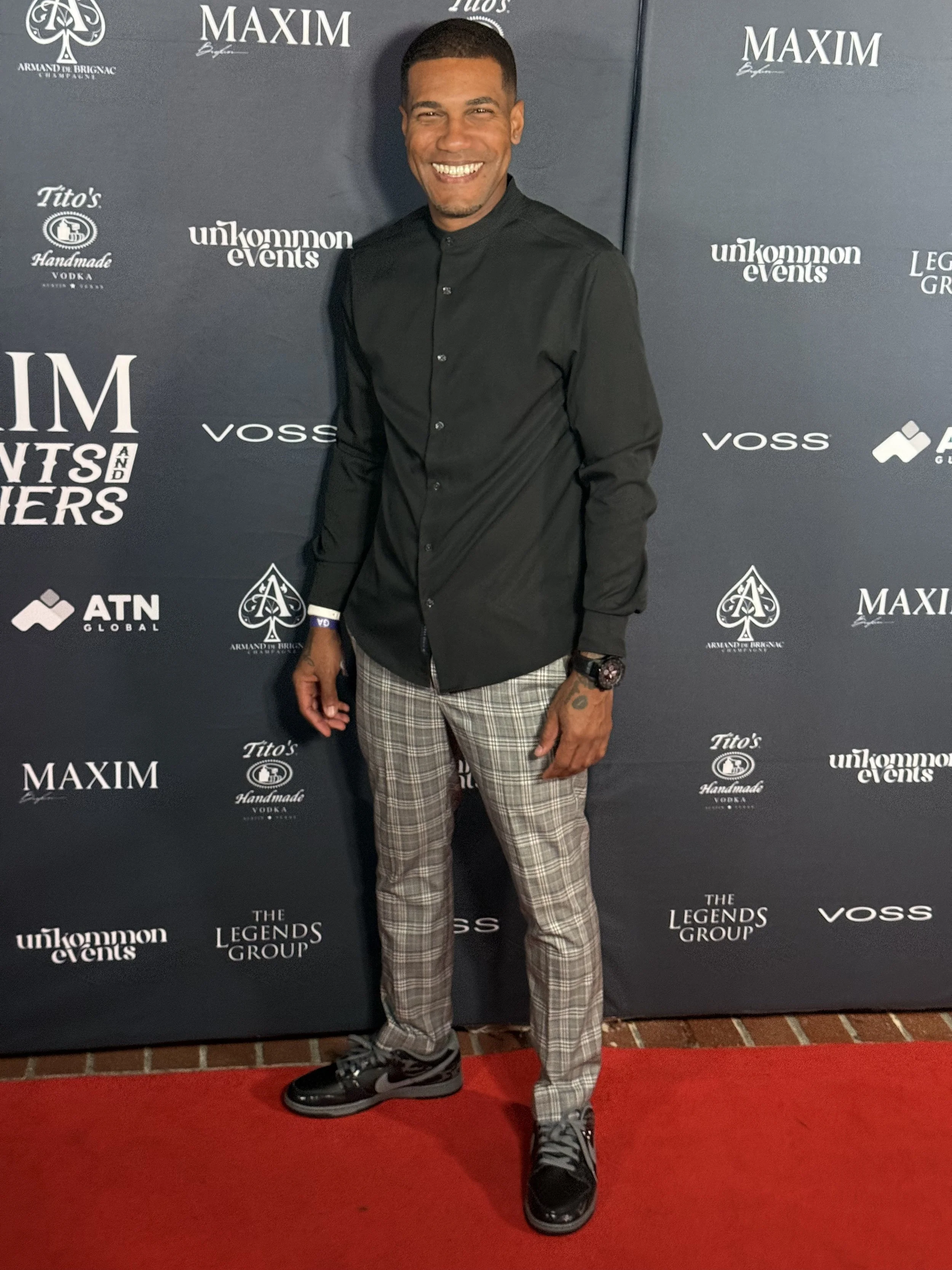 A man smiling on a red carpet at an event with a backdrop featuring logos including Maxim, unknown events, Tito's Handmade Vodka, The Legends Group, Voss, and ATN Global. He is wearing a black shirt, plaid pants, and black sneakers.