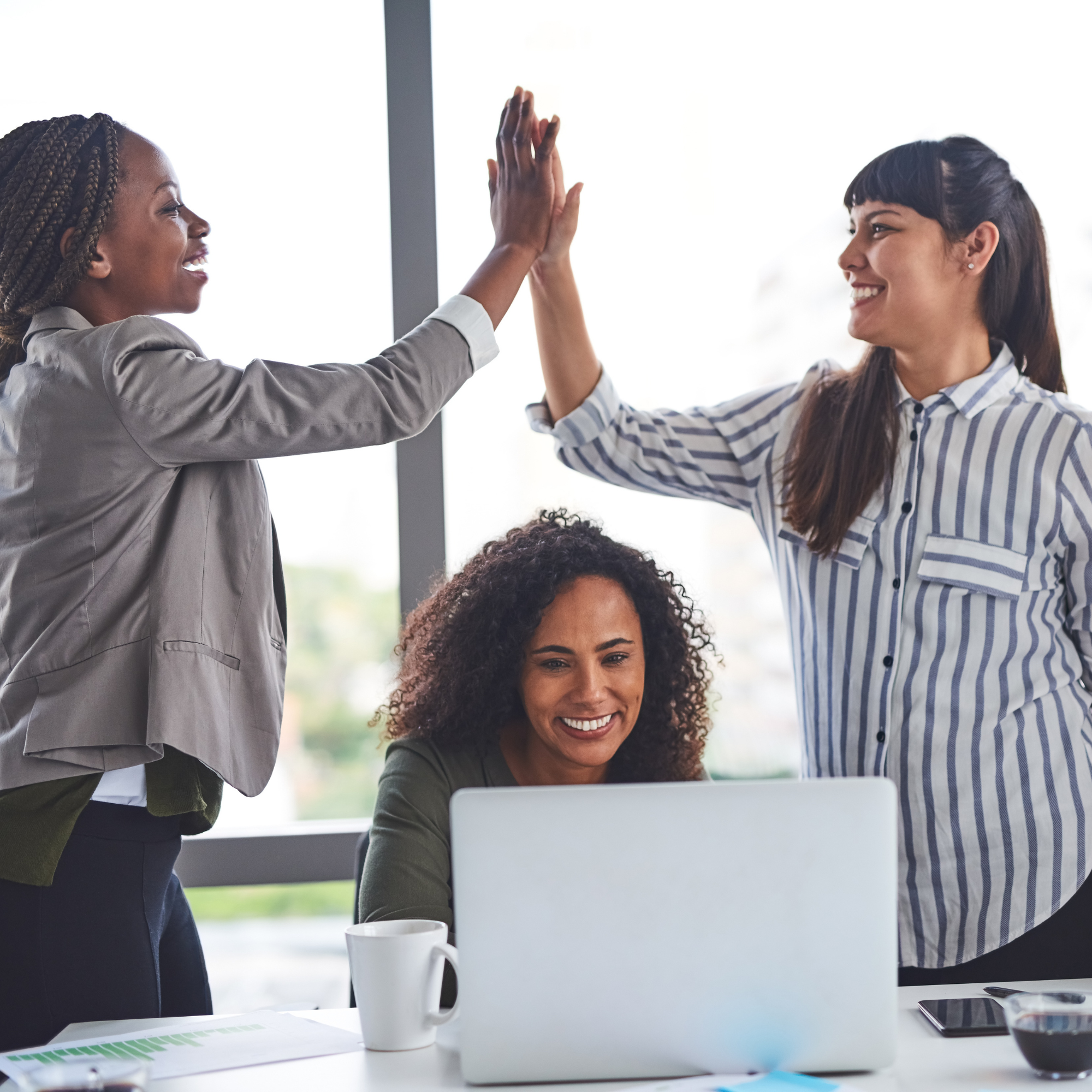 Two women giving each other a high five in an office environment, with one woman sitting at a desk and the other standing nearby, both smiling.