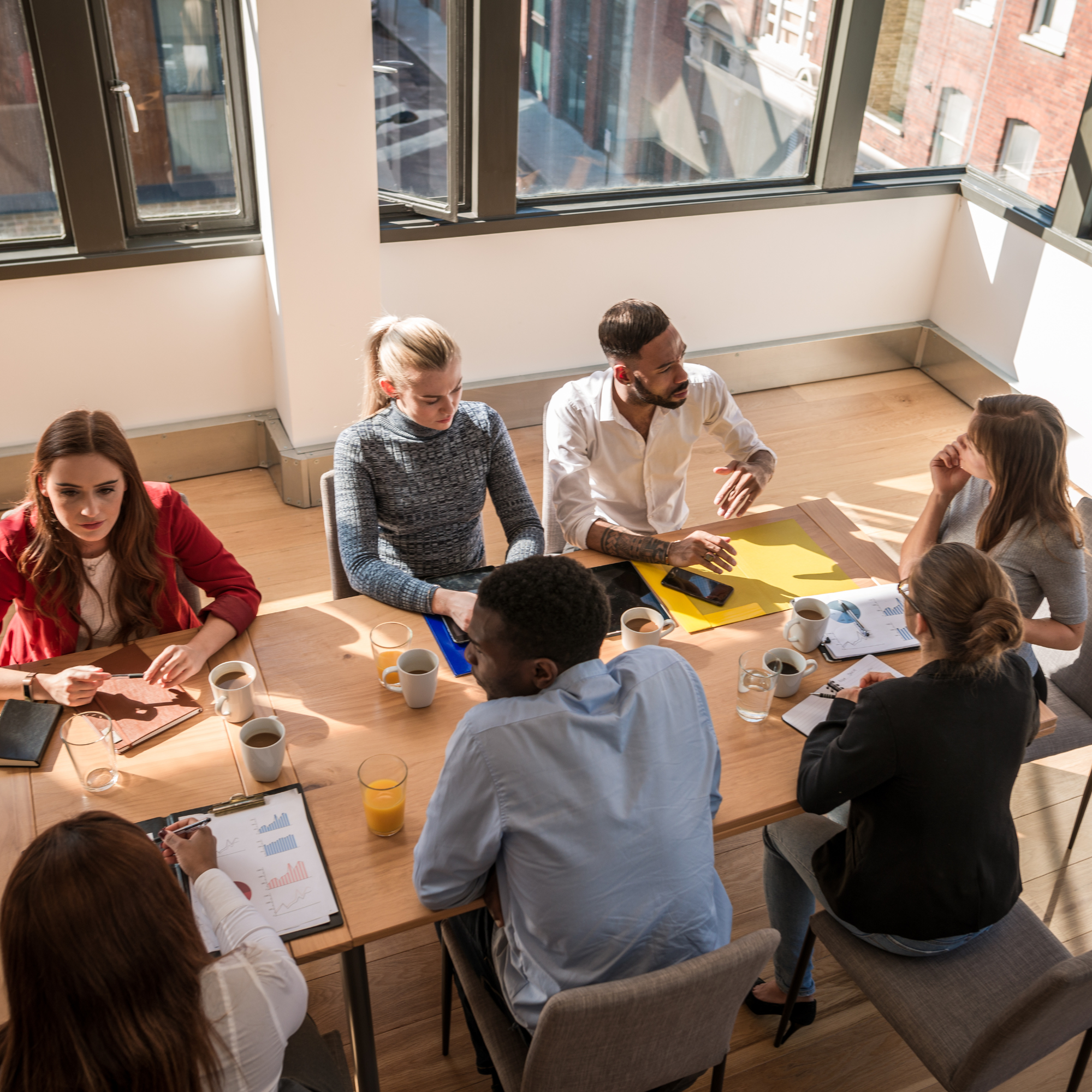A group of diverse business people having a meeting in a bright conference room with large windows showing city buildings outside. They are seated around a wooden table with cups of coffee and various documents, some engaging in conversation or looking at papers.