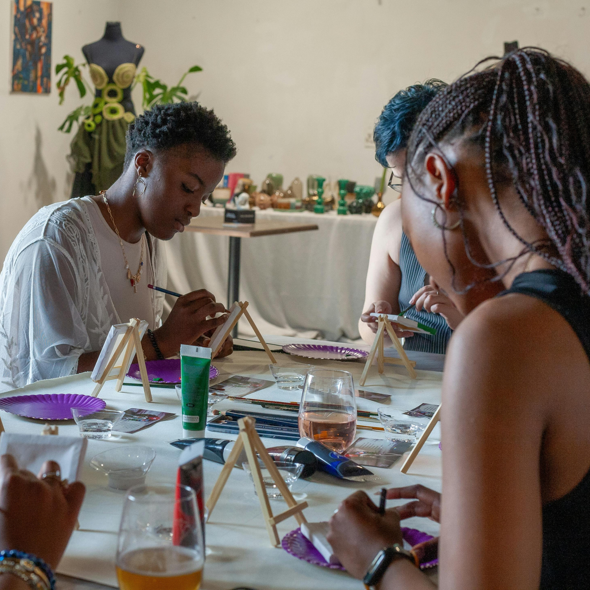 People painting miniature models at a table decorated with purple plates, cups, and art supplies, with an artwork of a black mannequin with green swirls in the background.