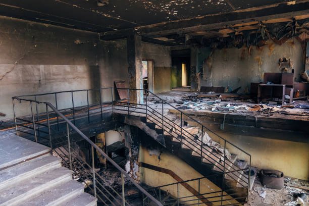 Interior of a burned-out building with charred walls, debris scattered across the floor, and a staircase leading to an upper level.