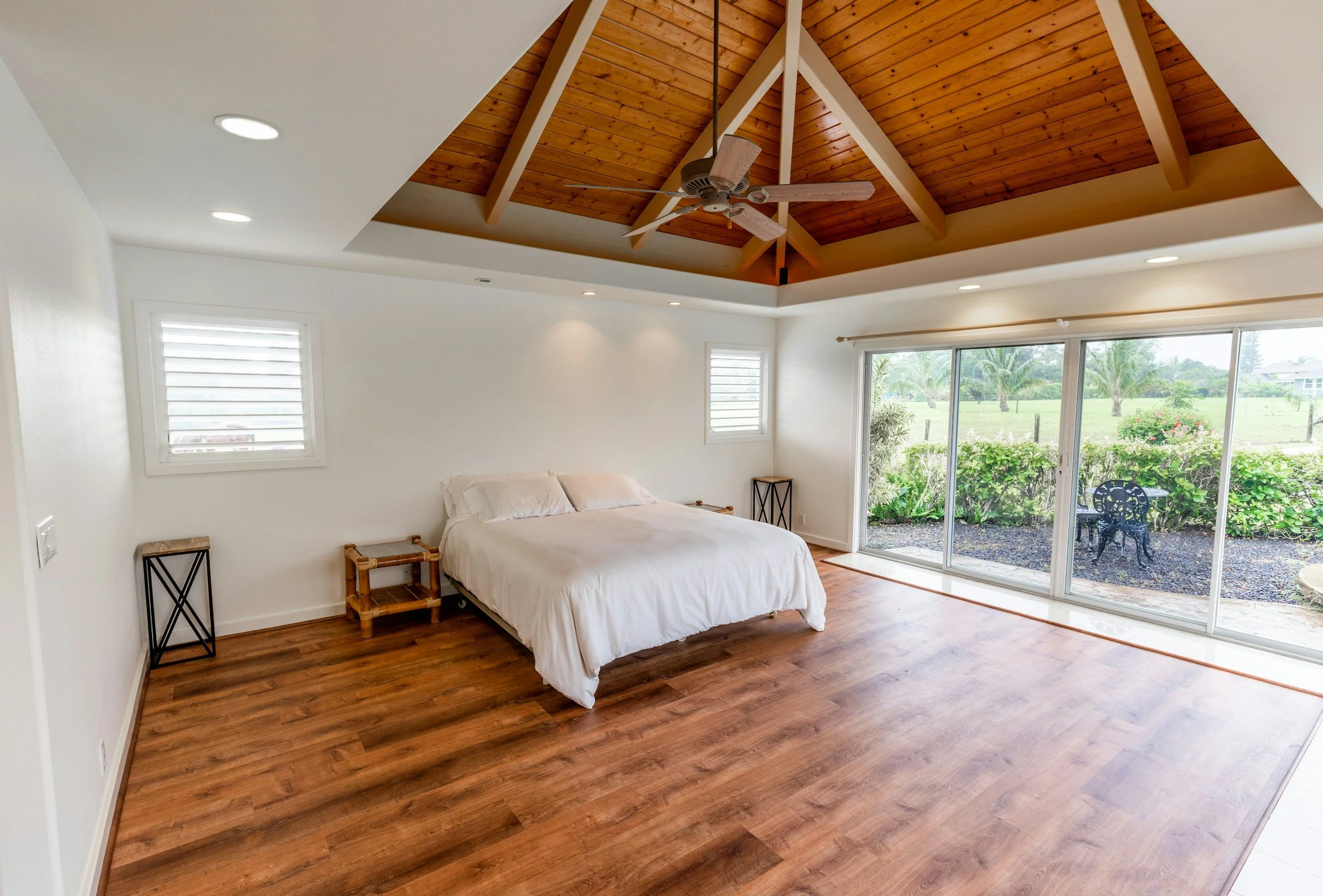 Bedroom with a wooden vaulted ceiling, ceiling fan, white walls, two small windows, large sliding glass door leading to patio, bed with white linens, small nightstand and decorative chair outside